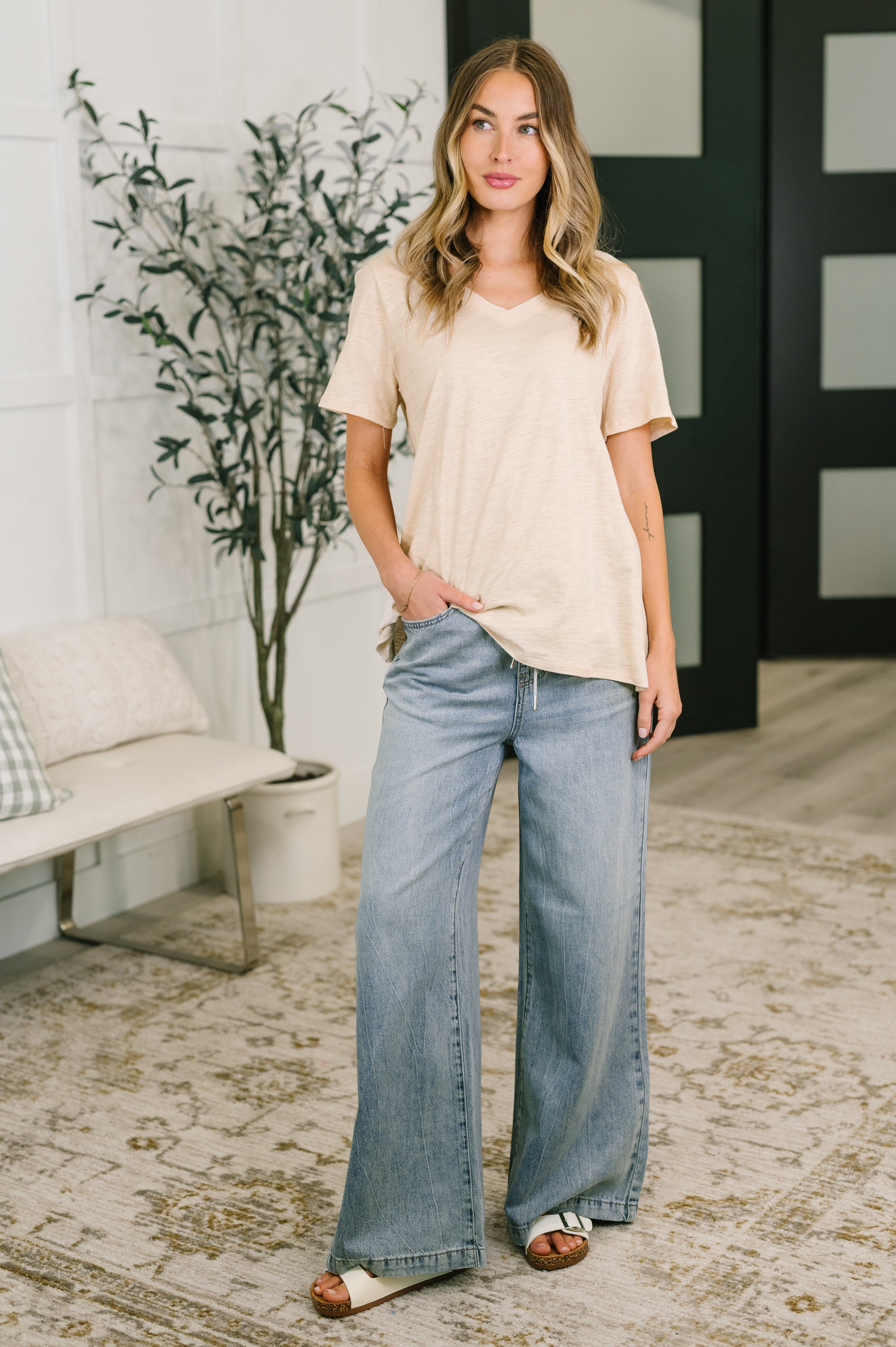 Woman in oatmeal V-neck tee and wide-leg light-wash jeans standing casually in a bright modern room