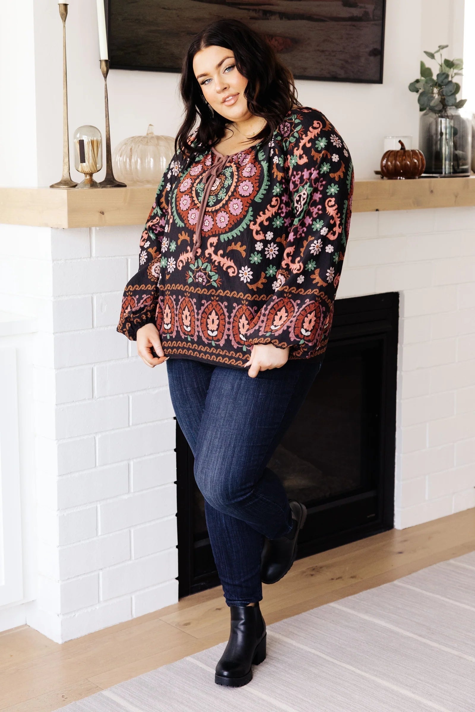 Woman indoors wearing a black I Feel That Peasant Blouse with colorful intricate floral patterns paired with dark jeans and black boots