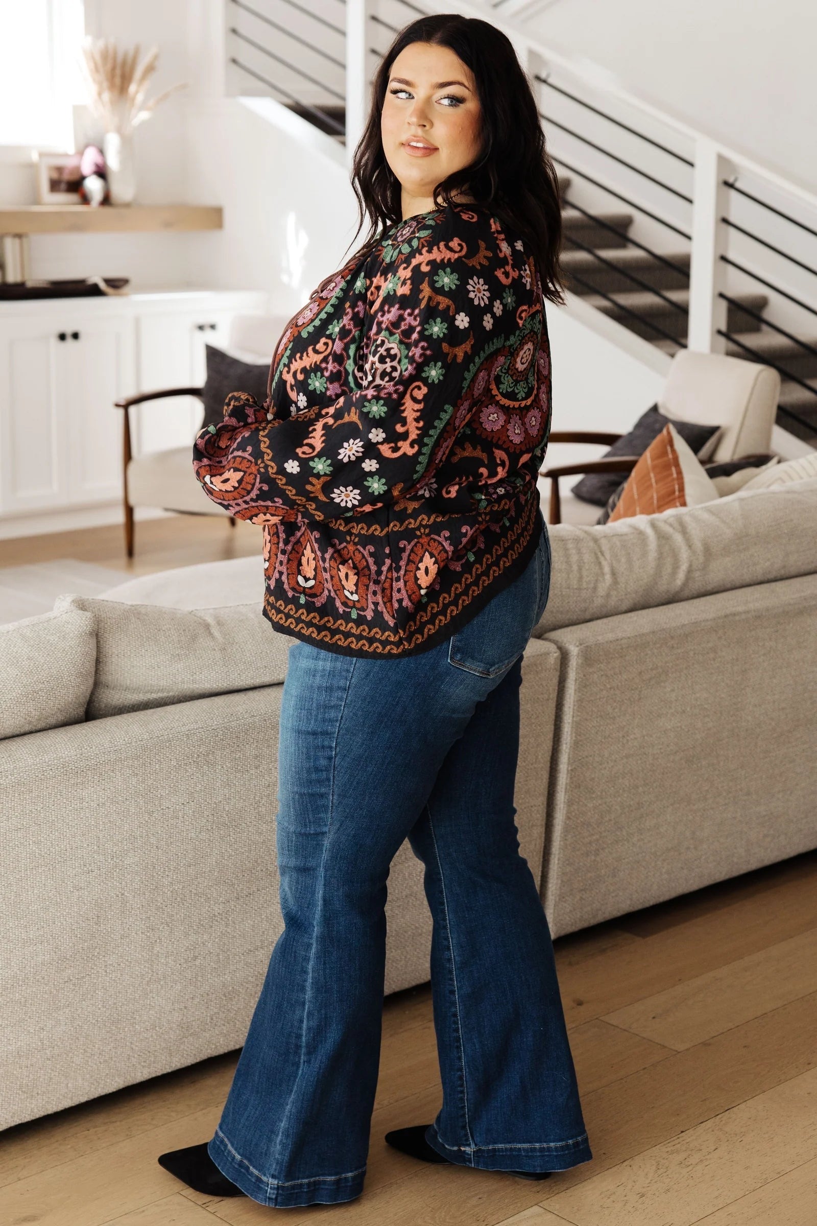 Plus size woman indoors wearing black I Feel That Peasant Blouse with multicolored paisley patterns paired with blue flared jeans