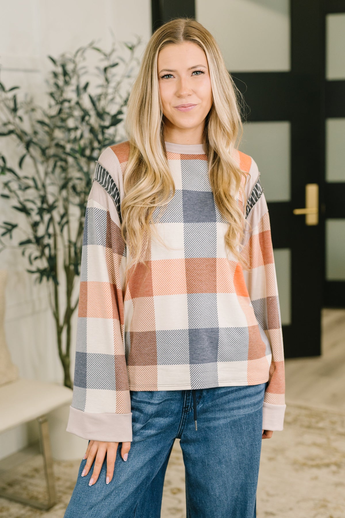 Woman models an orange plaid relaxed top with long sleeves and blue jeans in a bright modern entryway