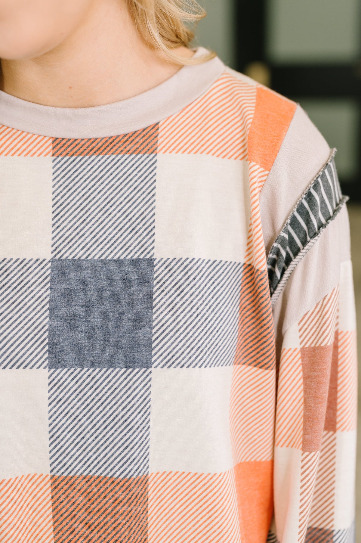 Close-up of a woman wearing an orange plaid relaxed top with a banded round neckline and contrasting seam detail on the sleeve