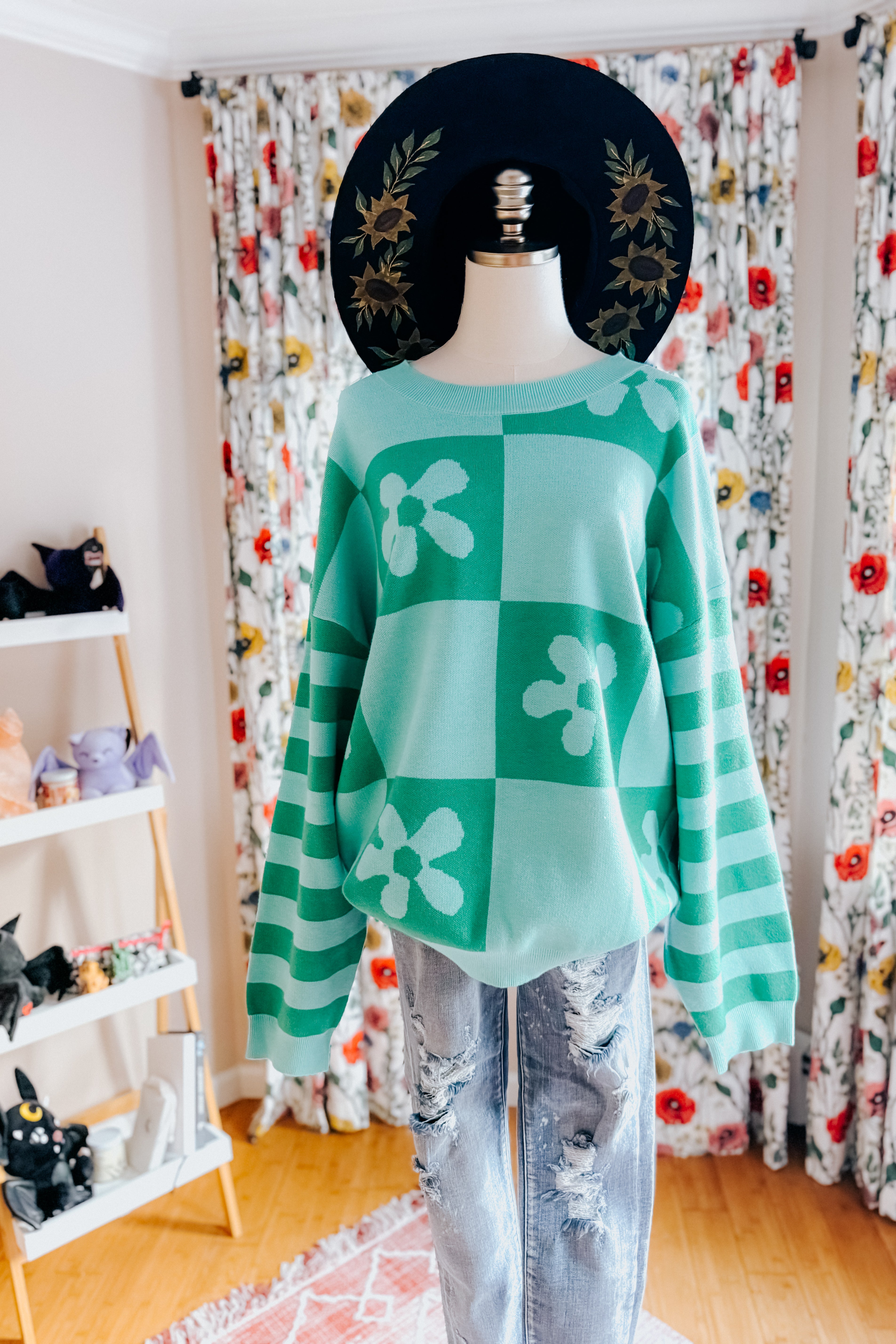 A dress form wearing a checkered floral print mint sweater, blue jeans and a painted wide brim hat in an indoor setting 