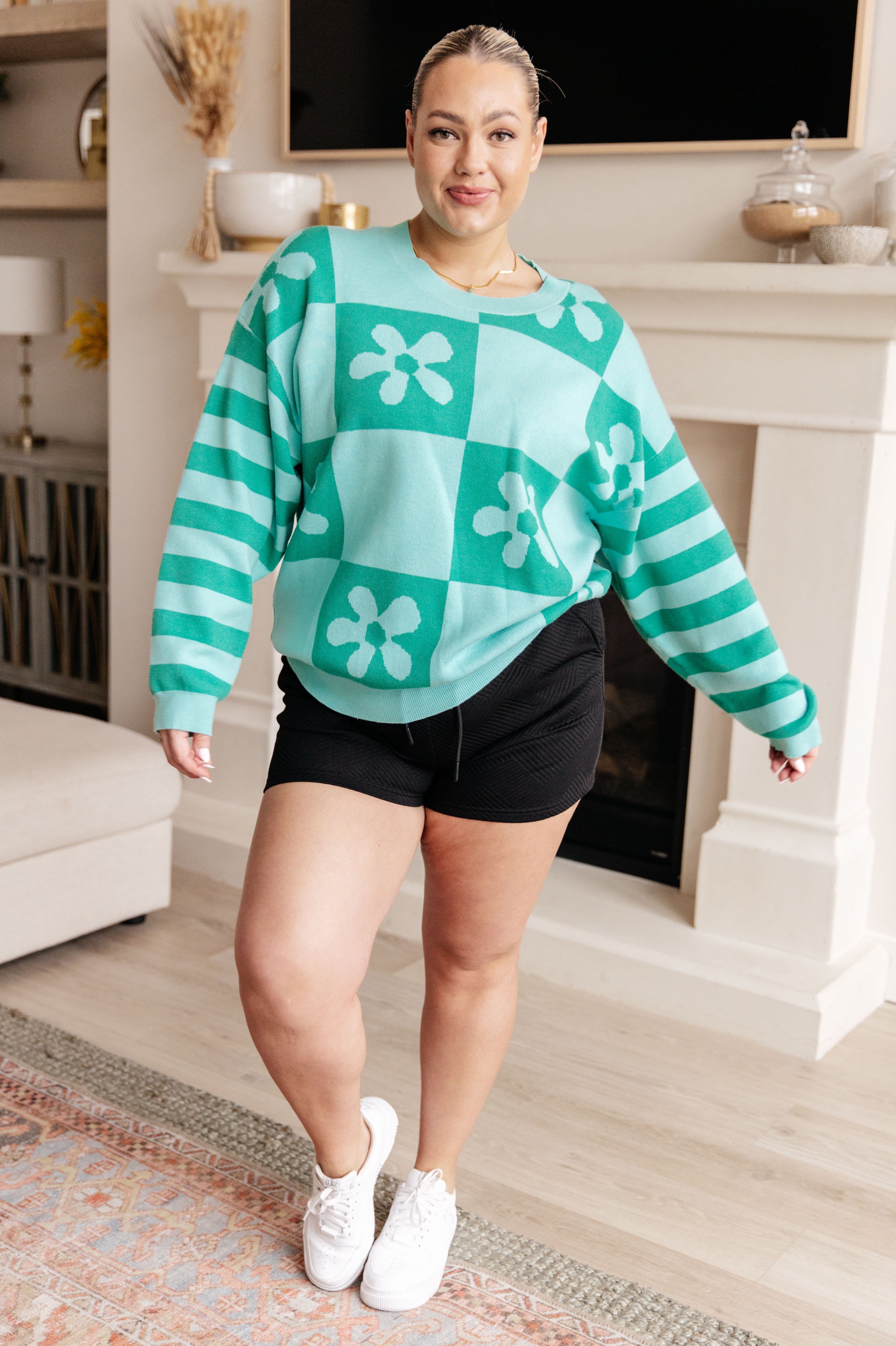 Woman wearing a mint floral checkered print sweater, black shorts and white sneakers in a living room.