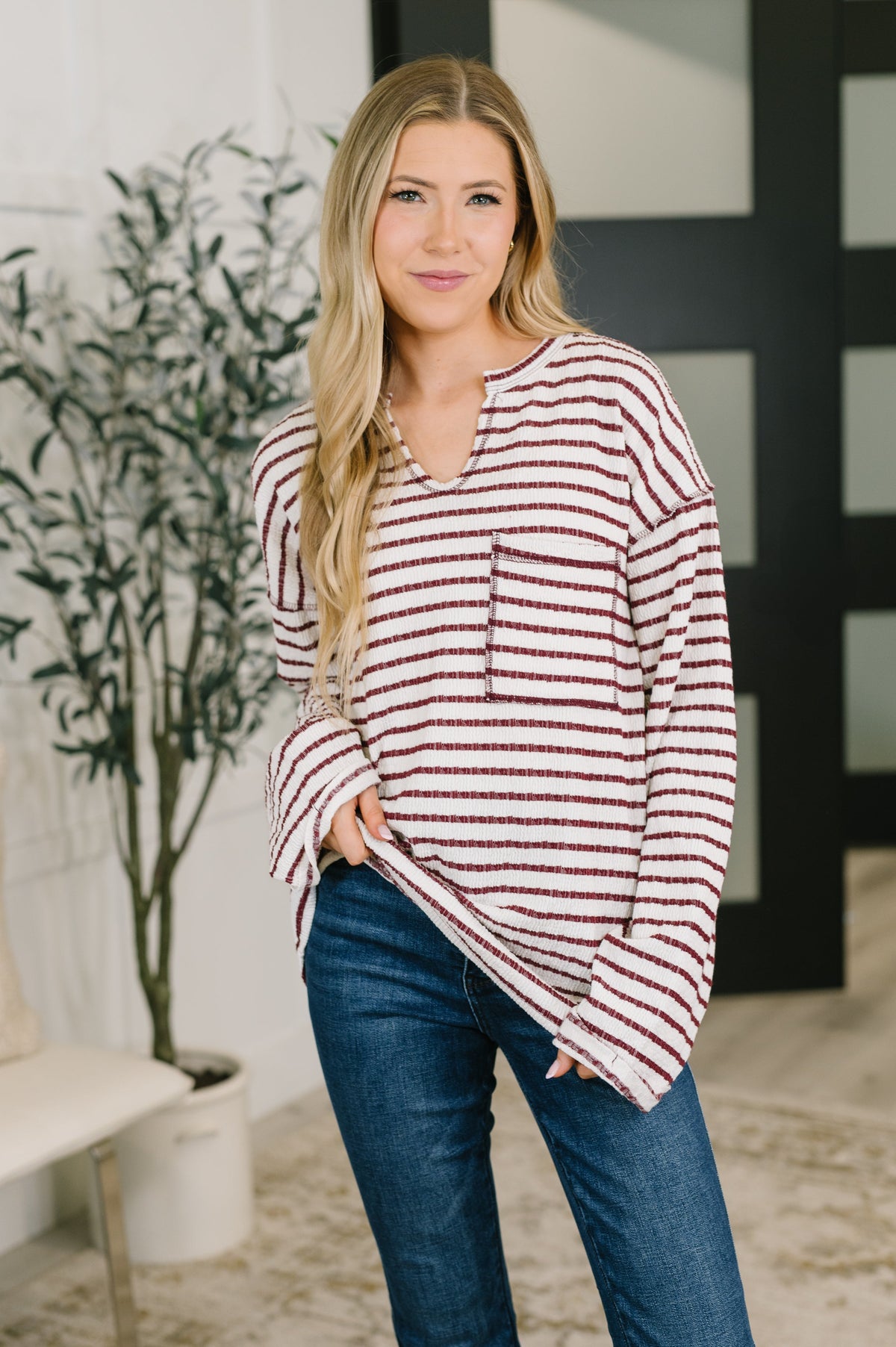 Woman wearing a cream and burgundy striped relaxed knit long sleeve top with split neckline and pocket paired with jeans indoors