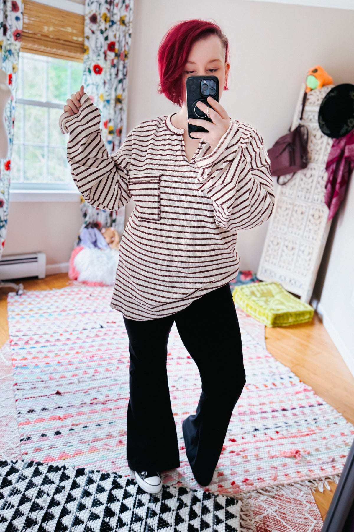 Red haired woman taking a bedroom mirror selfie in a cream striped knit top with split neckline black pants and sneakers
