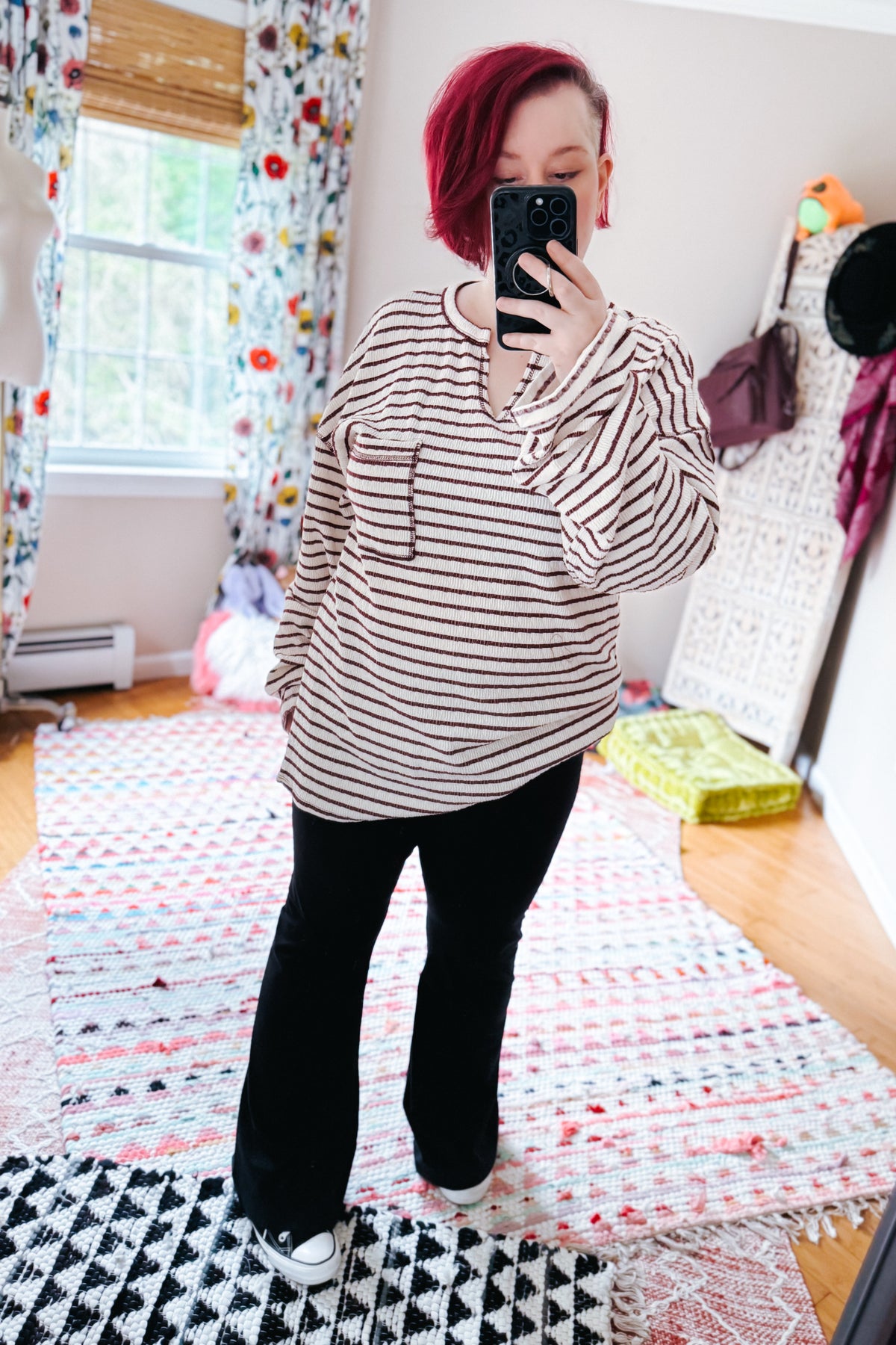 Red haired woman taking a bedroom mirror selfie in a cream striped knit long sleeve top with split neckline and black pants