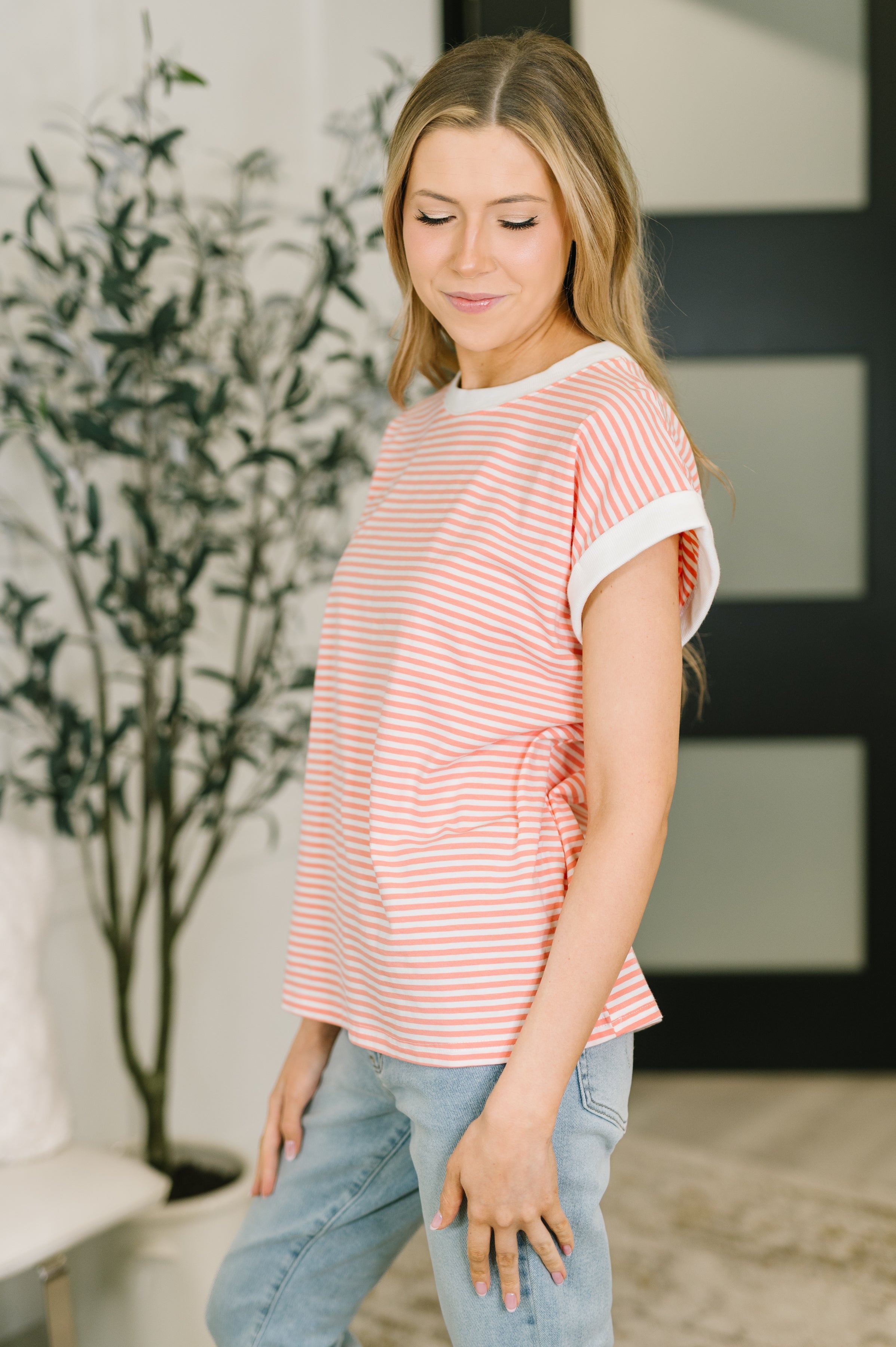 Woman wearing coral and white horizontal striped short sleeve top with white trim, standing indoors near plant and black door