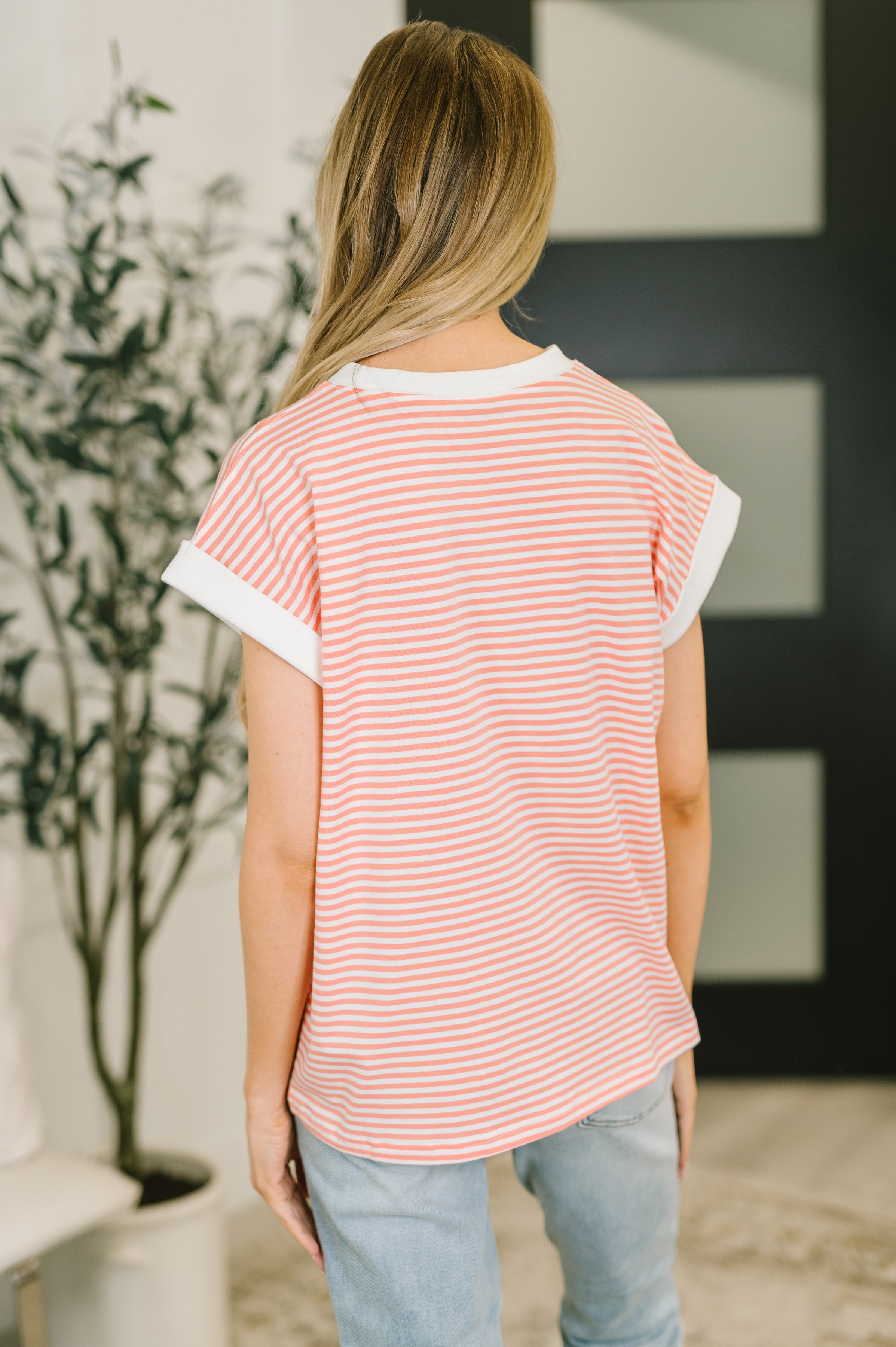 Woman wearing a coral and white striped short sleeve top with rolled cuffs standing indoors with back to camera