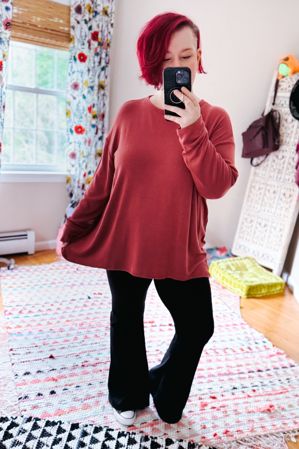 Woman wearing a cranberry long sleeve top with dropped shoulders styled with black pants and white shoes in a colorful bedroom mirror selfie