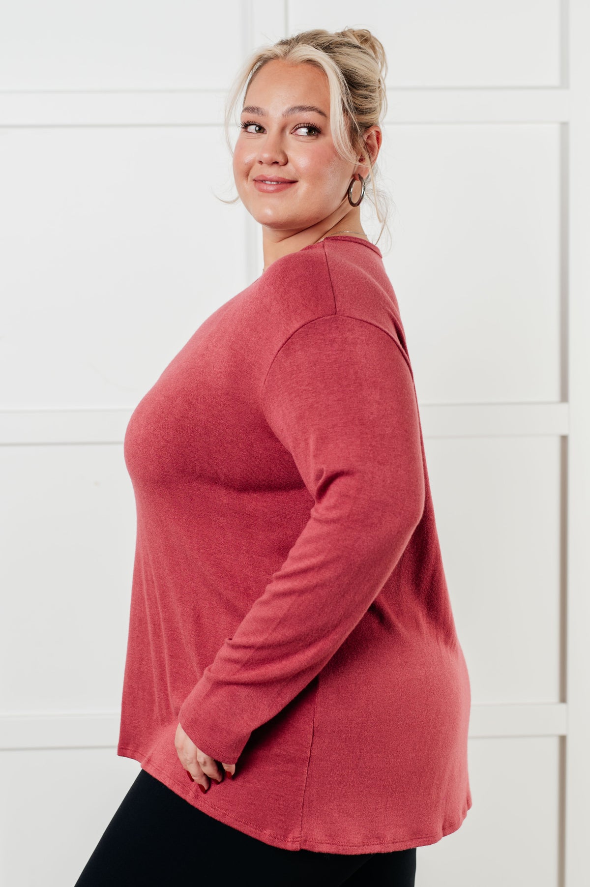Woman modeling a cranberry long sleeve top with a relaxed drop shoulder fit and curved hem against a bright paneled wall