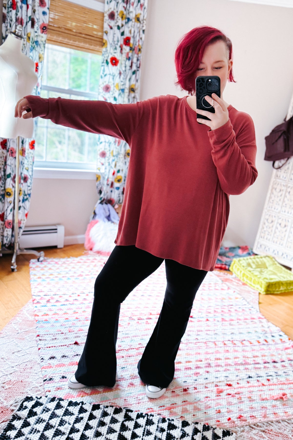 Woman taking a mirror selfie in a cranberry long sleeve top with dropped shoulders styled with black pants and white shoes indoors