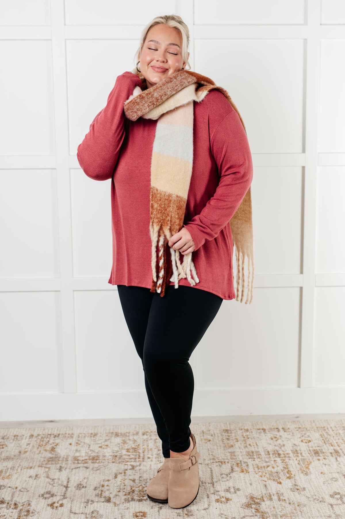 Smiling woman wearing a cranberry long sleeve top with a relaxed fit styled with a fringed scarf black leggings and tan ankle boots indoors