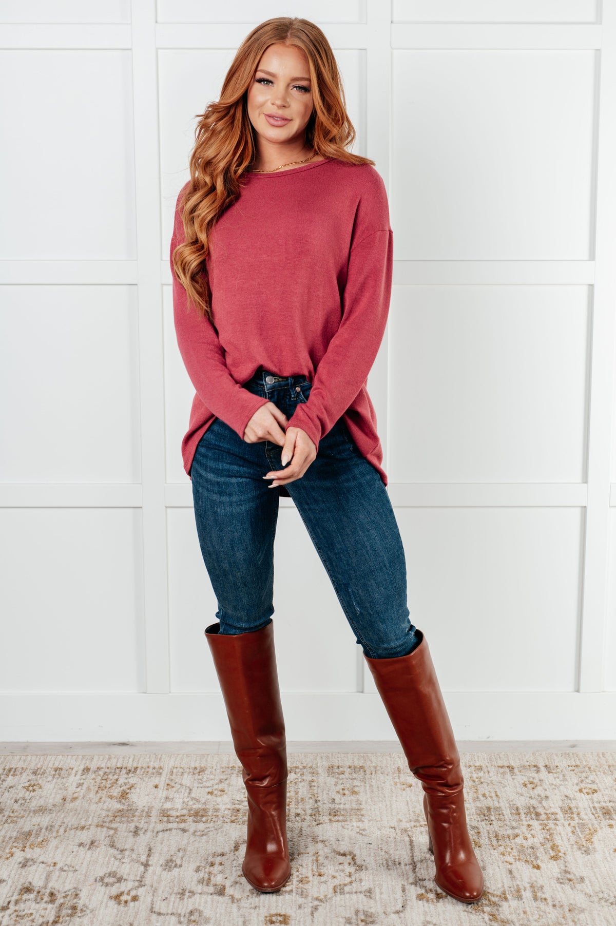 Woman modeling a cranberry long sleeve top with relaxed drop shoulders, dark jeans, and tall brown boots against a white paneled wall