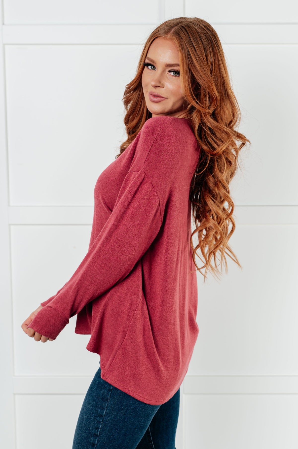 Woman modeling a cranberry long sleeve top with relaxed drop shoulders and a curved hem paired with dark jeans against a white wall