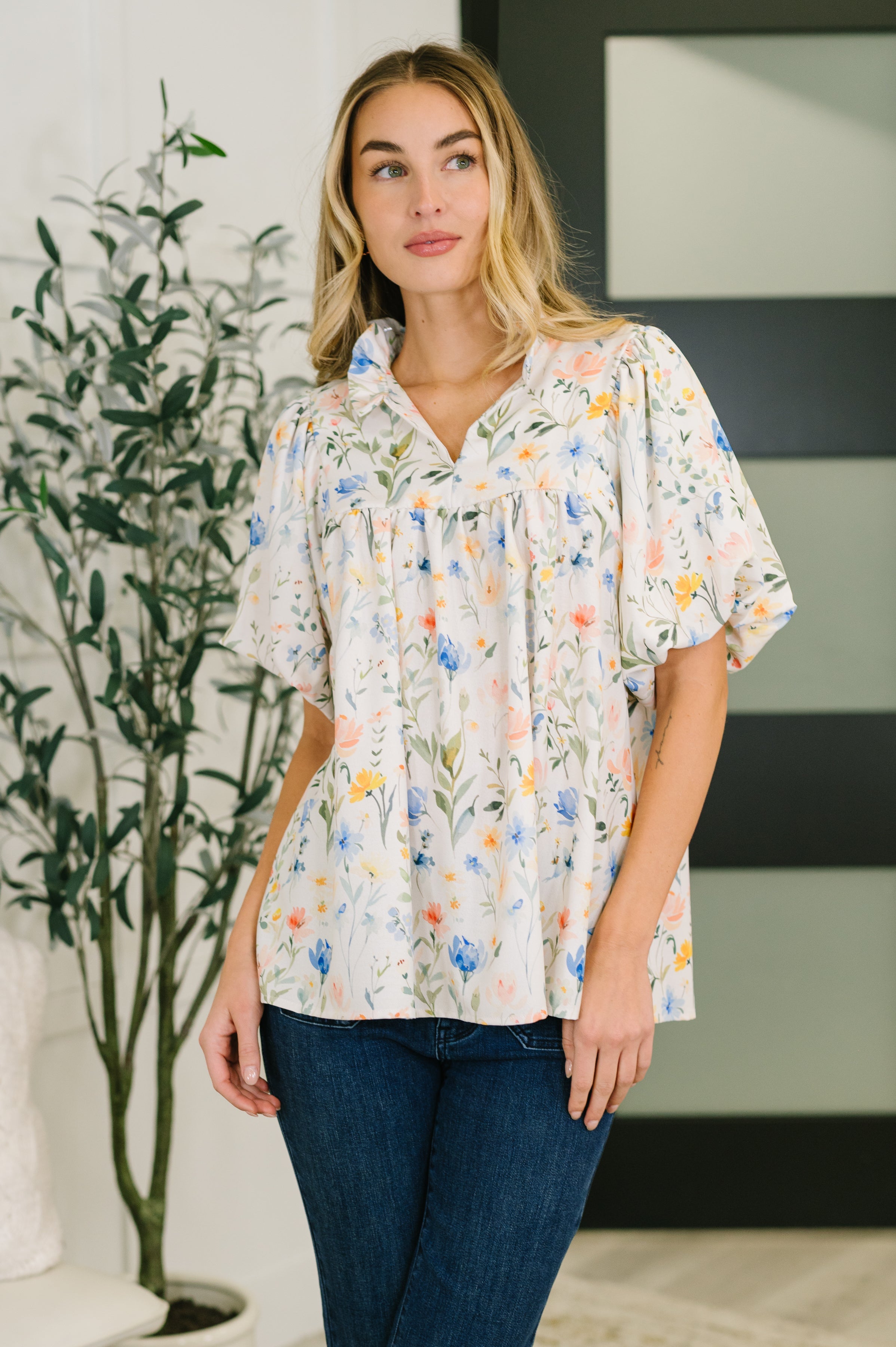 Woman wearing cream floral notch neck blouse with ruffle details and puff sleeves styled with dark jeans in bright space