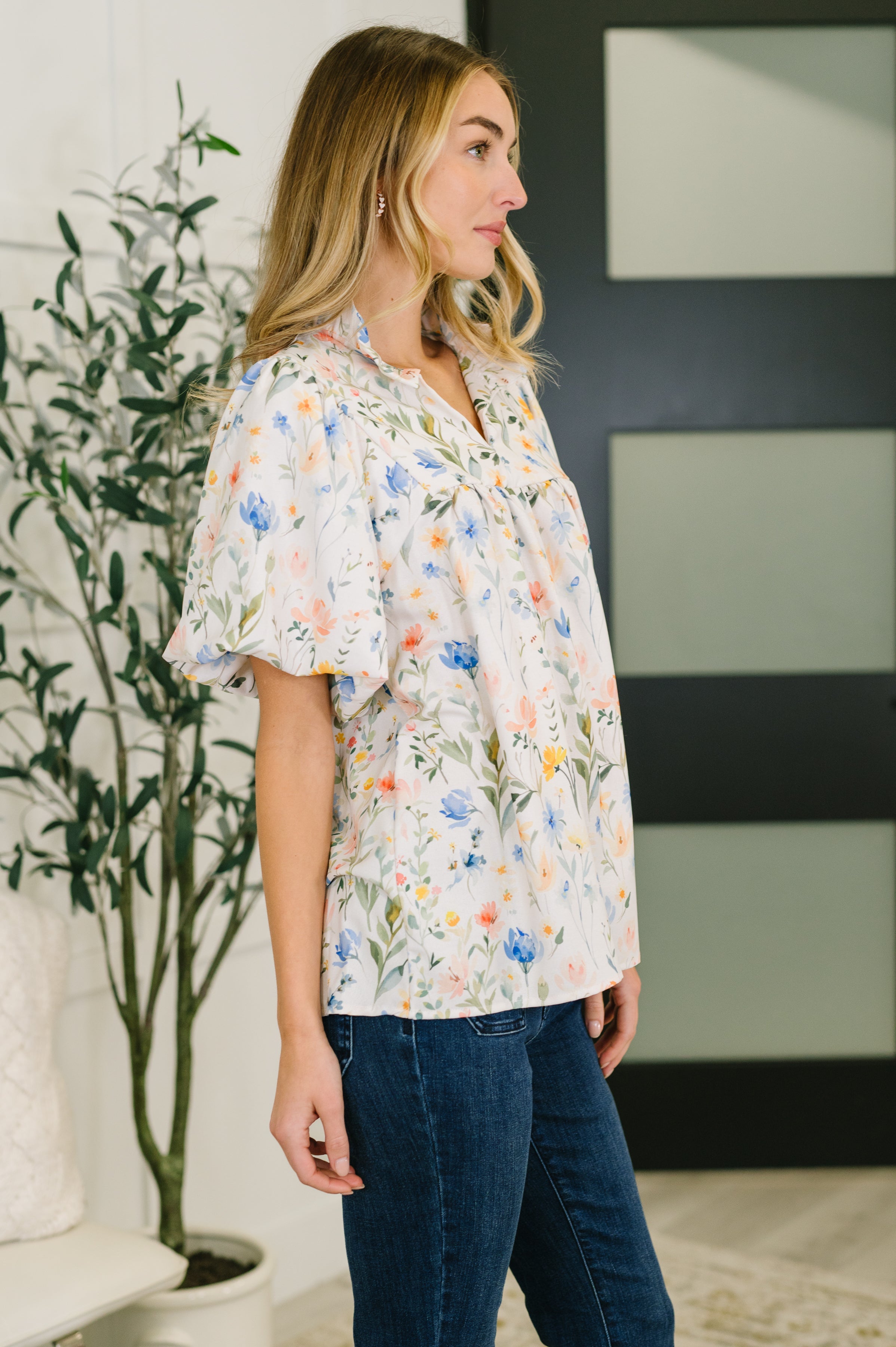 Woman wearing cream floral blouse with notch neck and puff sleeves paired with dark blue jeans in bright eclectic space