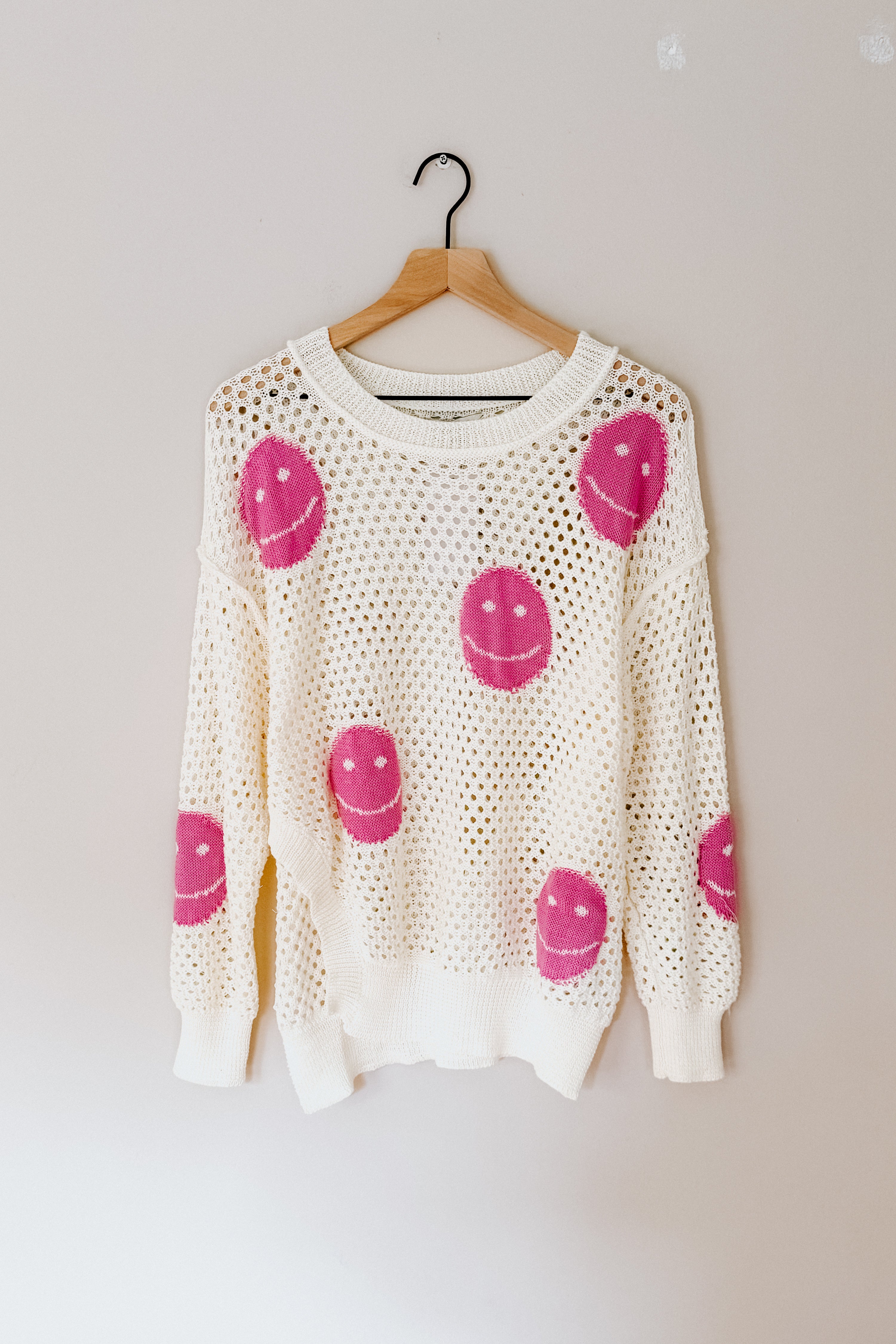 Beige open-knit top with bright pink smiley face designs hanging on wooden hanger against plain wall