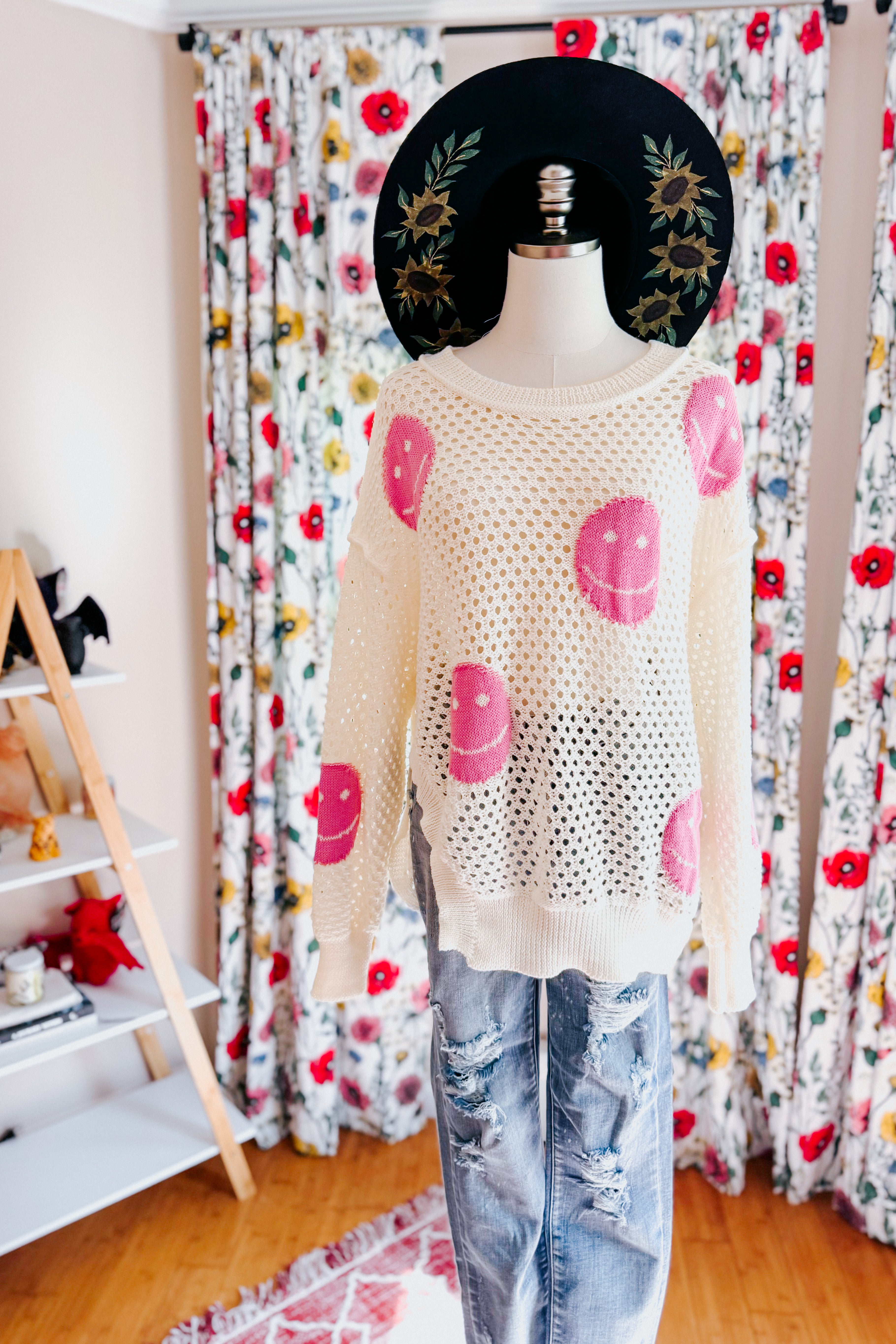 Mannequin displaying beige and pink In My Feelings Smiley Open Knit Top paired with distressed jeans and black floral wide-brim hat indoors