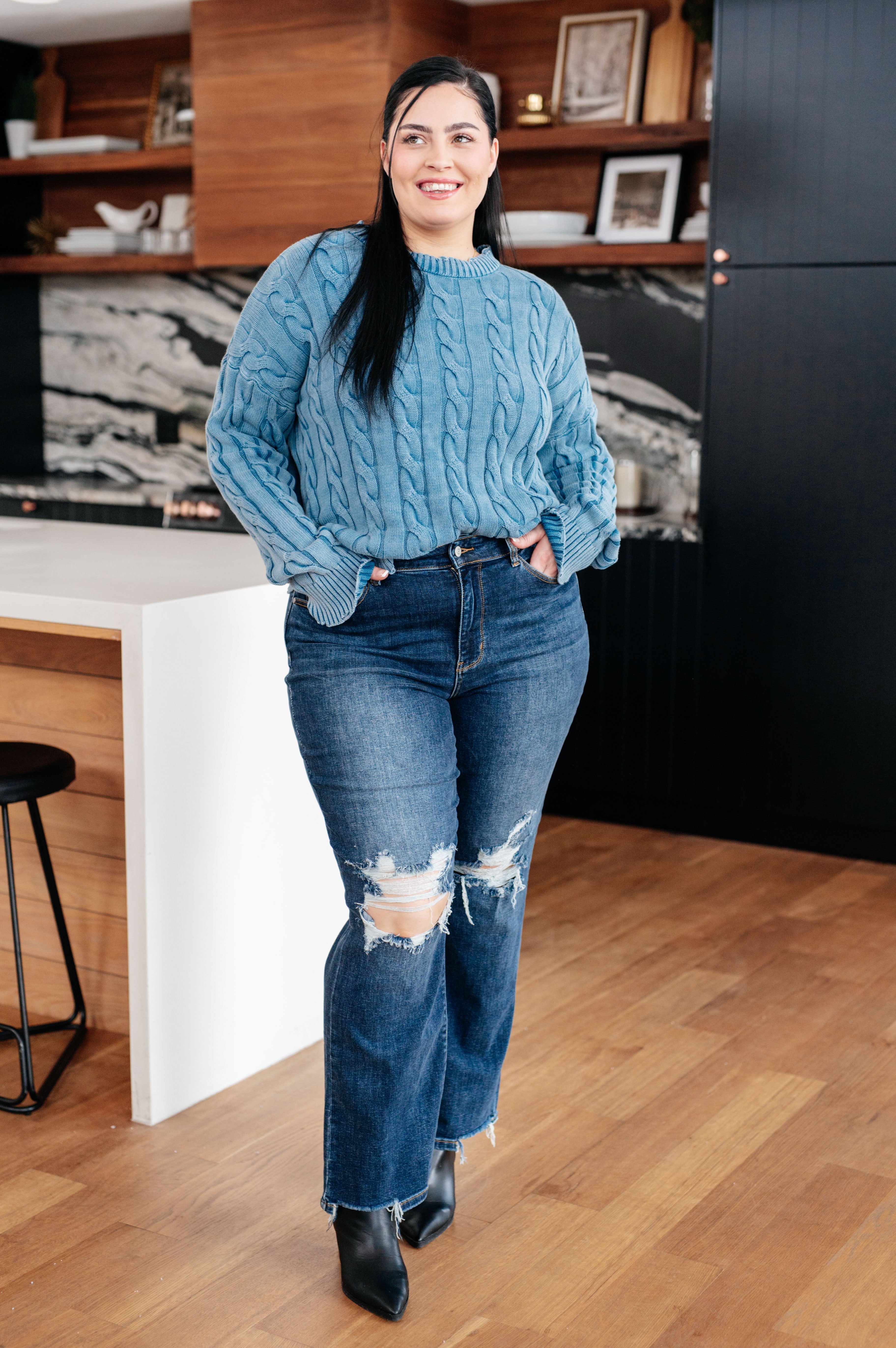 Smiling woman wearing blue In the Right Direction cable knit sweater with ribbed cuffs paired with distressed jeans and black boots indoors