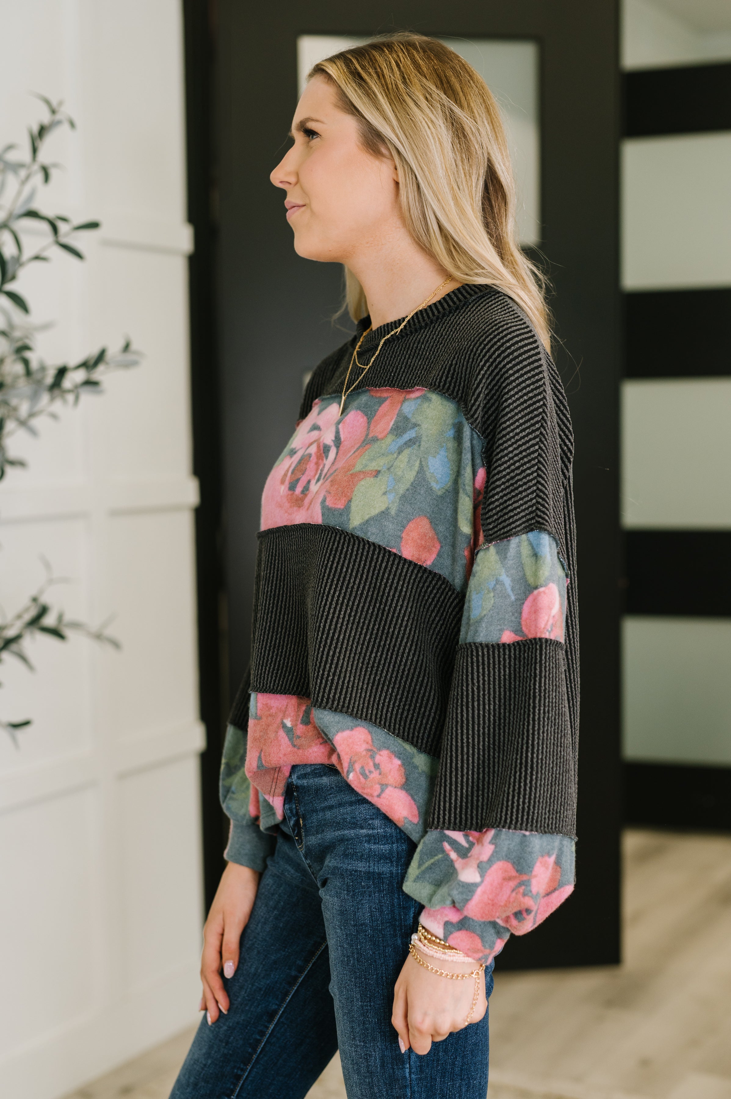 Woman wearing black ribbed pullover with pink and green floral panels paired with dark blue jeans indoors