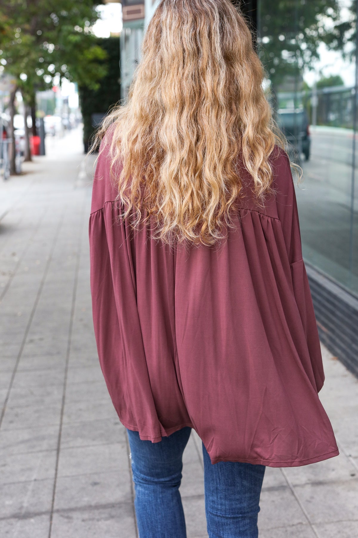 Woman walking on sidewalk wearing wine In This Groove modal knit top with loose fit and gathered back paired with blue jeans
