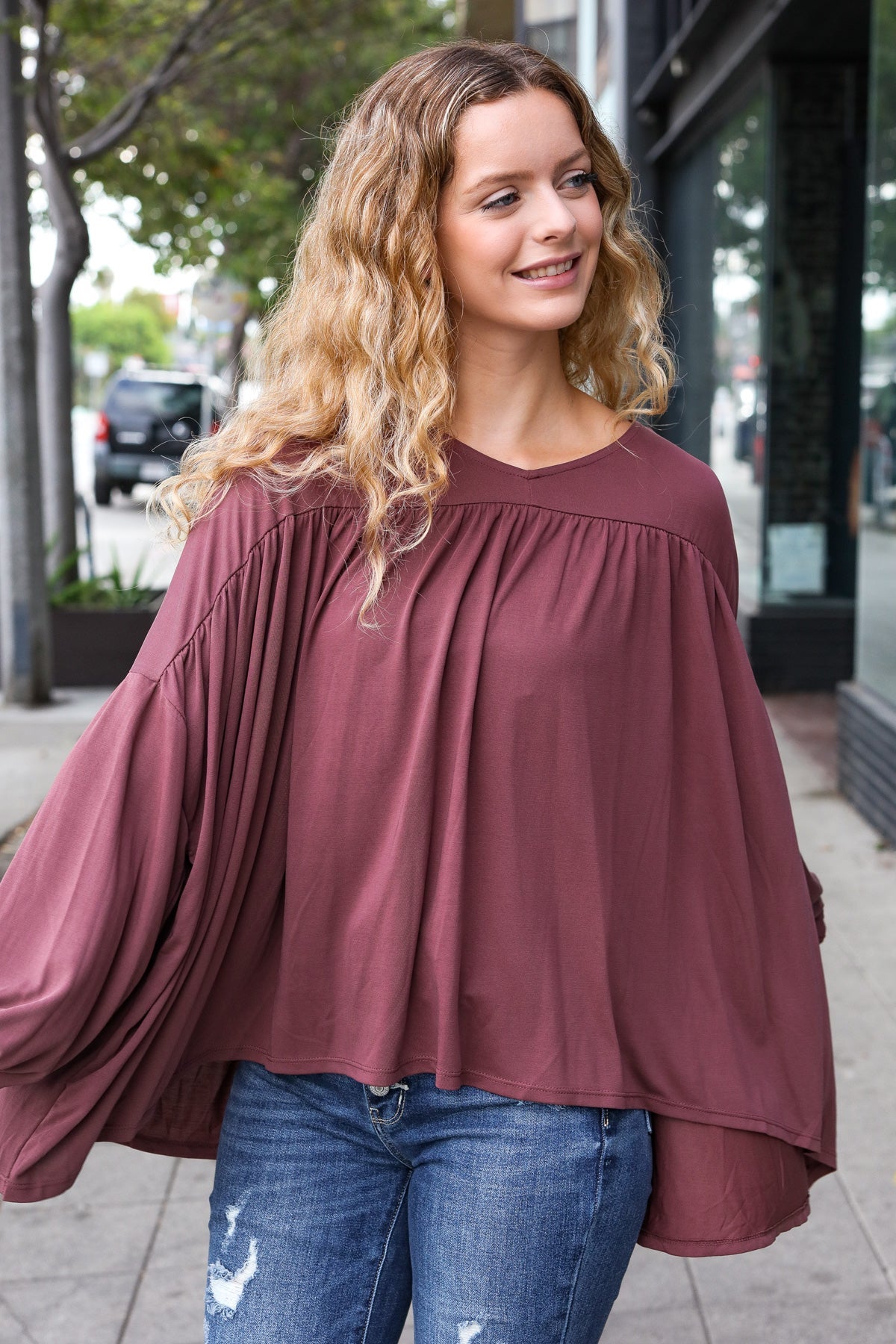 Woman smiling outdoors wearing wine In This Groove modal knit top with voluminous pleated sleeves and relaxed fit paired with blue jeans