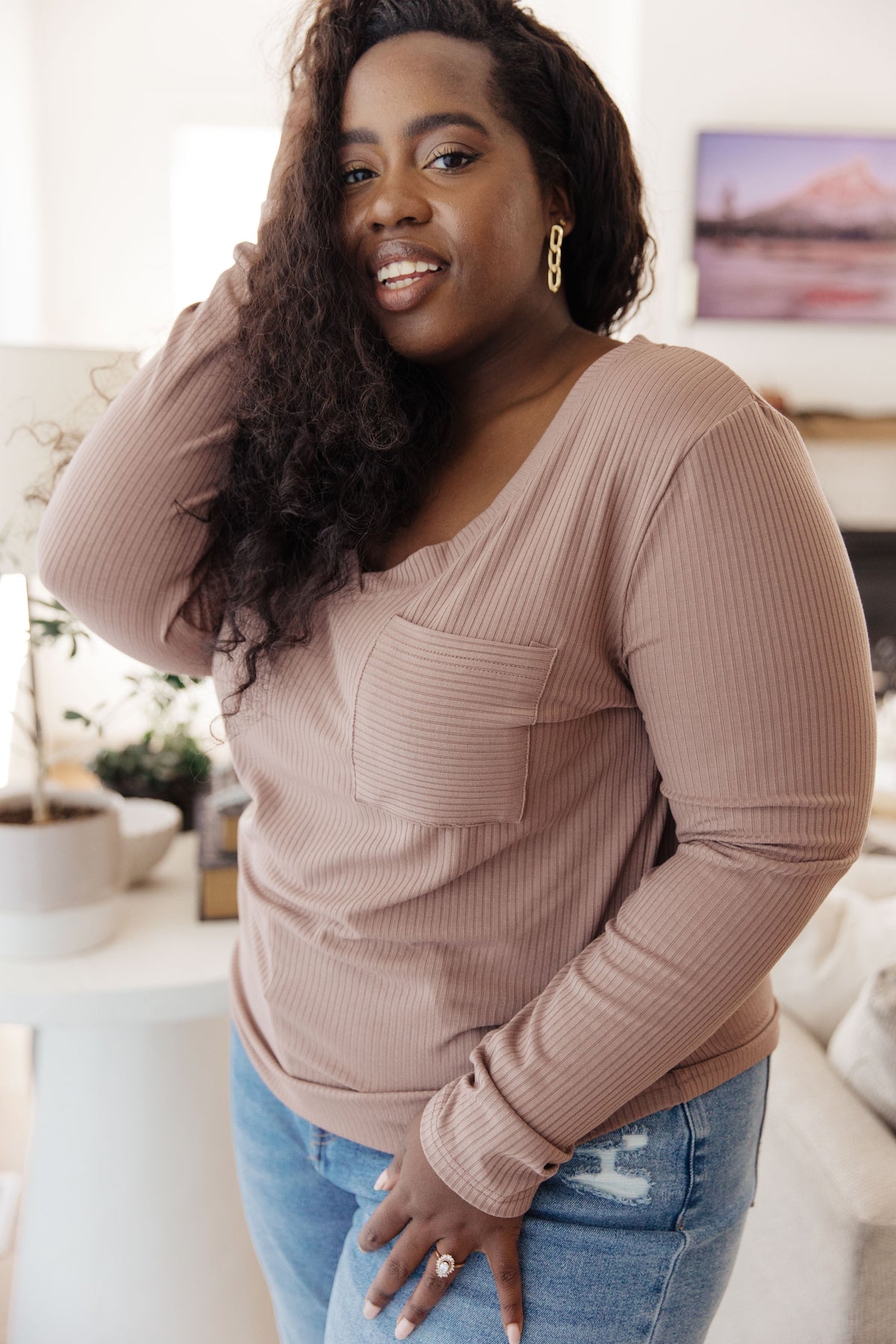 Smiling woman wearing a beige ribbed knit V-neck top with a patch pocket and long sleeves with blue distressed jeans indoors