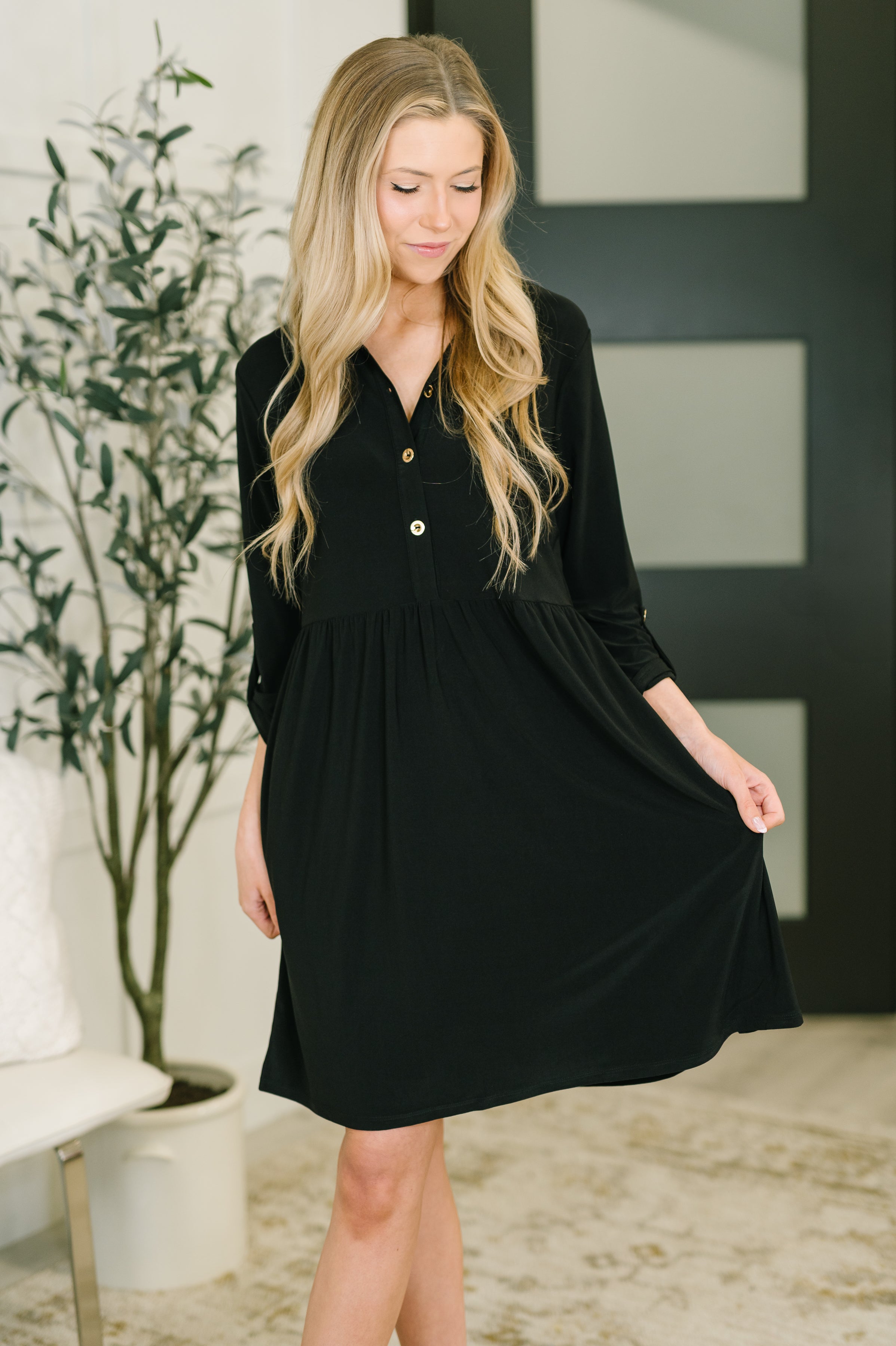 Woman indoors wearing black Inspire Collared Neck Dress with long sleeves and button front detail holding hem