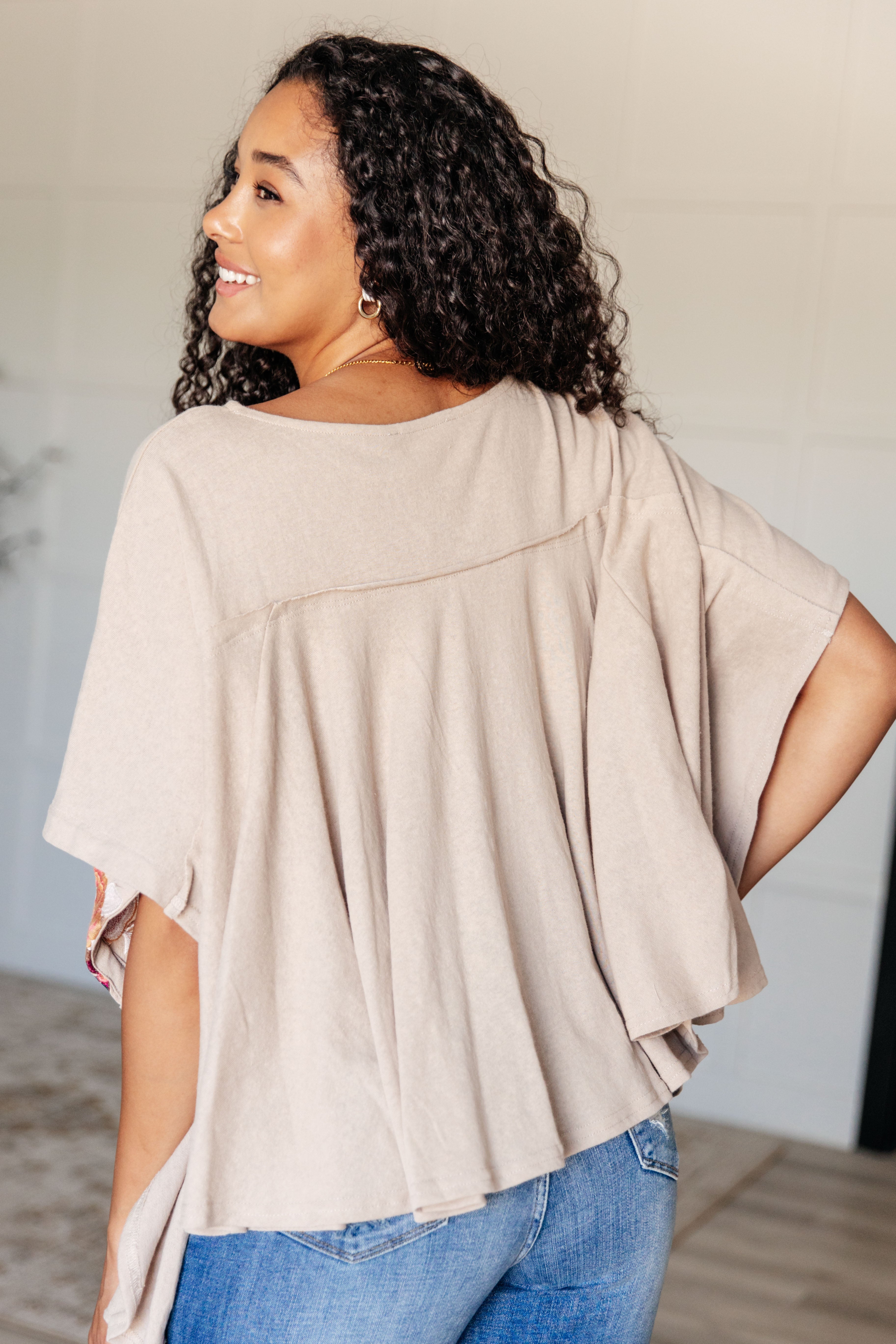 Woman with curly hair smiling wearing loose mocha Isabel tunic with flowing back pleats and short sleeves indoors