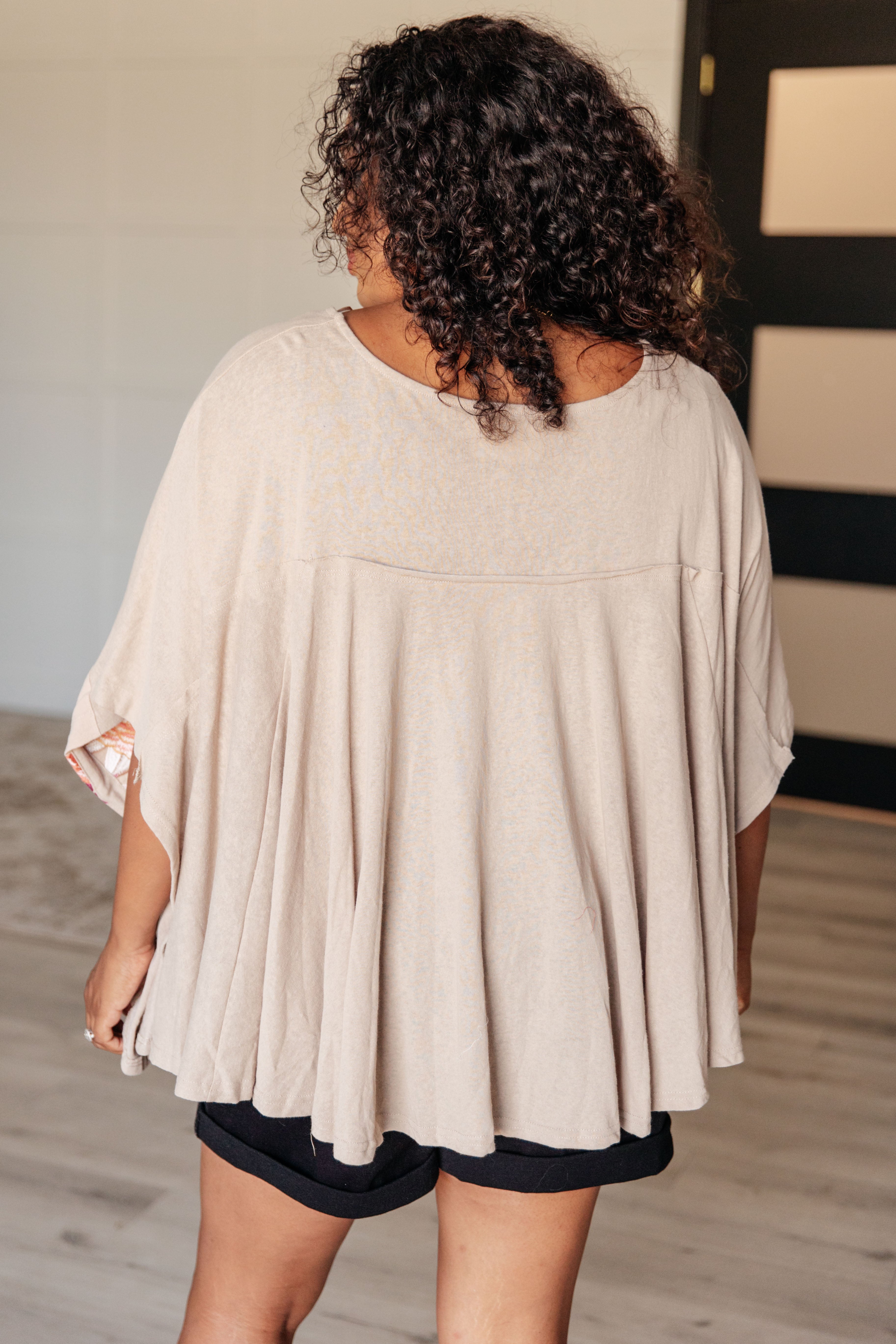 Curly-haired woman wearing loose-fitting mocha Isabel tunic seen from back with flowing pleated design and short sleeves