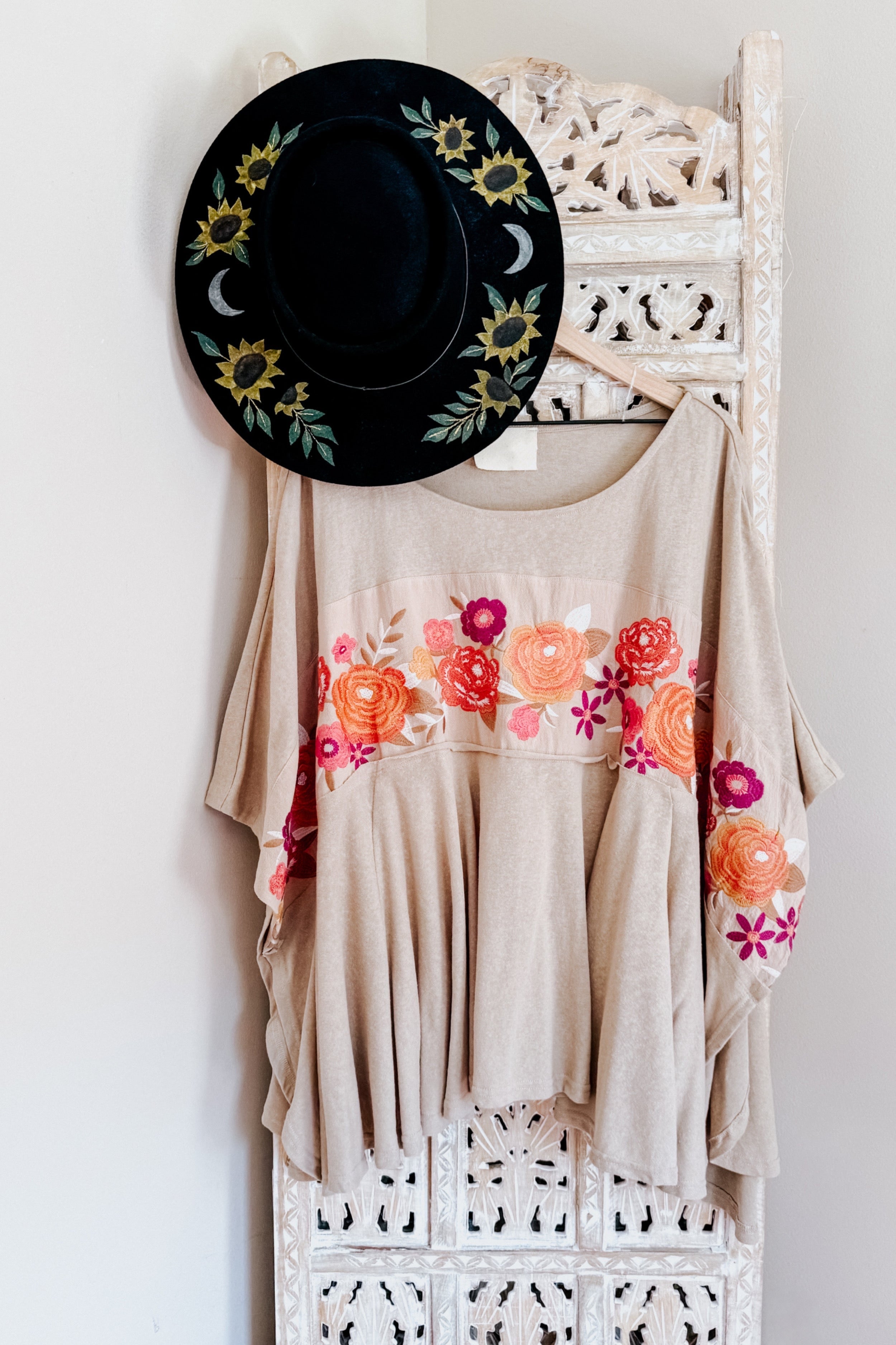Mocha Isabel tunic with vibrant floral embroidery in shades of orange and pink displayed on decorative white screen with black floral hat