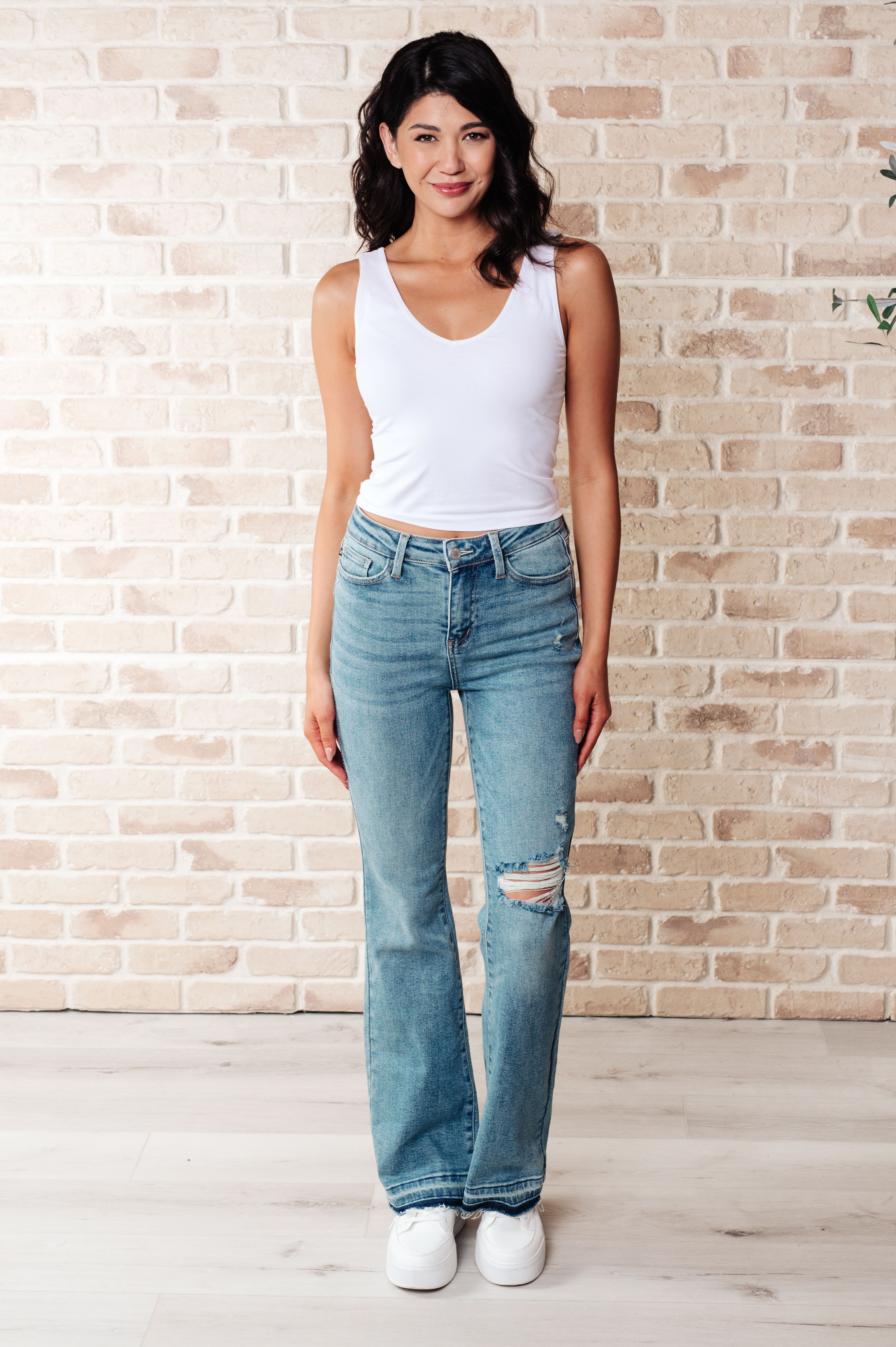 Woman wearing blue Isla mid rise bootcut jeans with distressed knee and released hems paired with white sneakers indoors