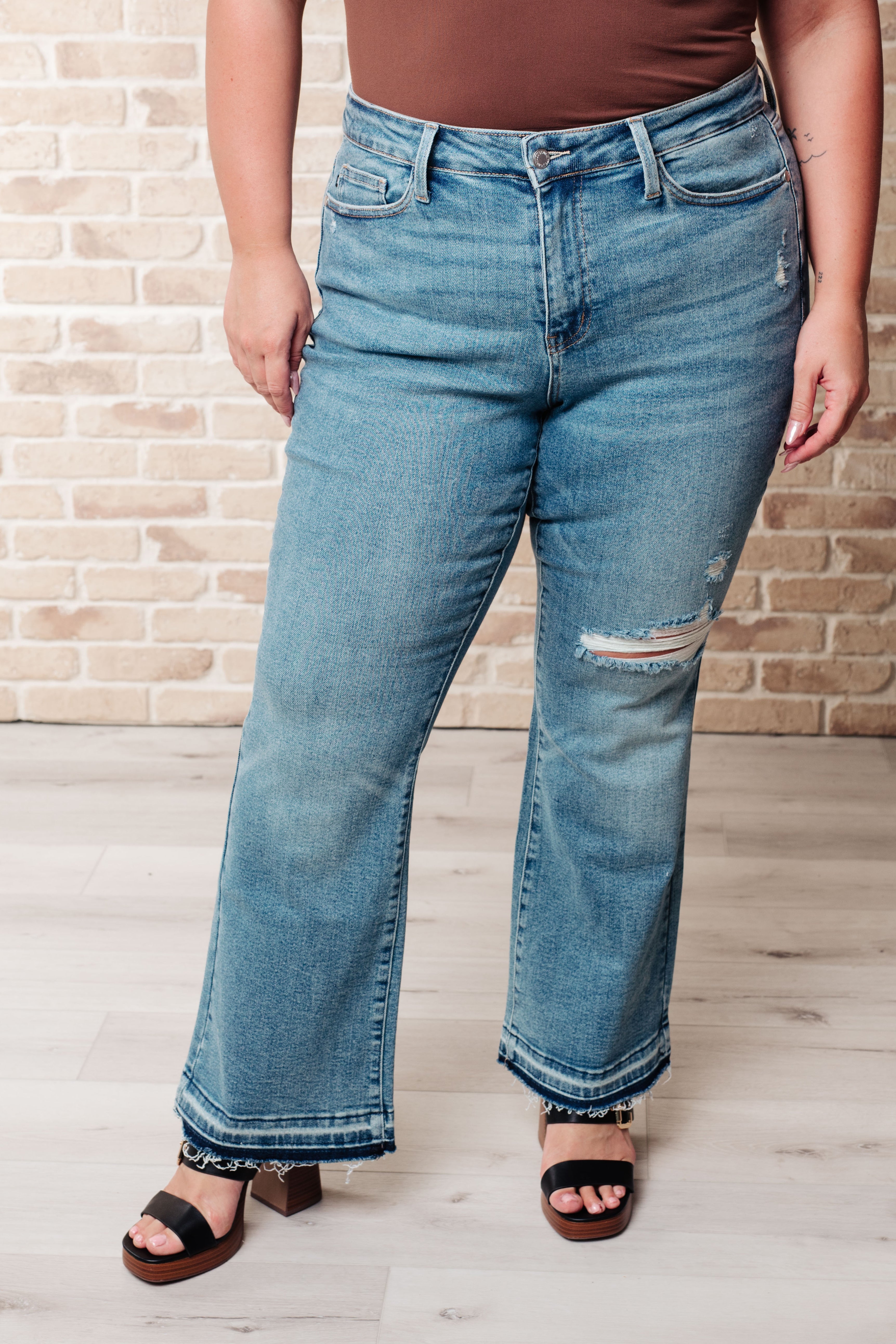 Front view of person wearing blue Isla mid rise bootcut jeans with distressed knee and raw released hems paired with black sandals indoors