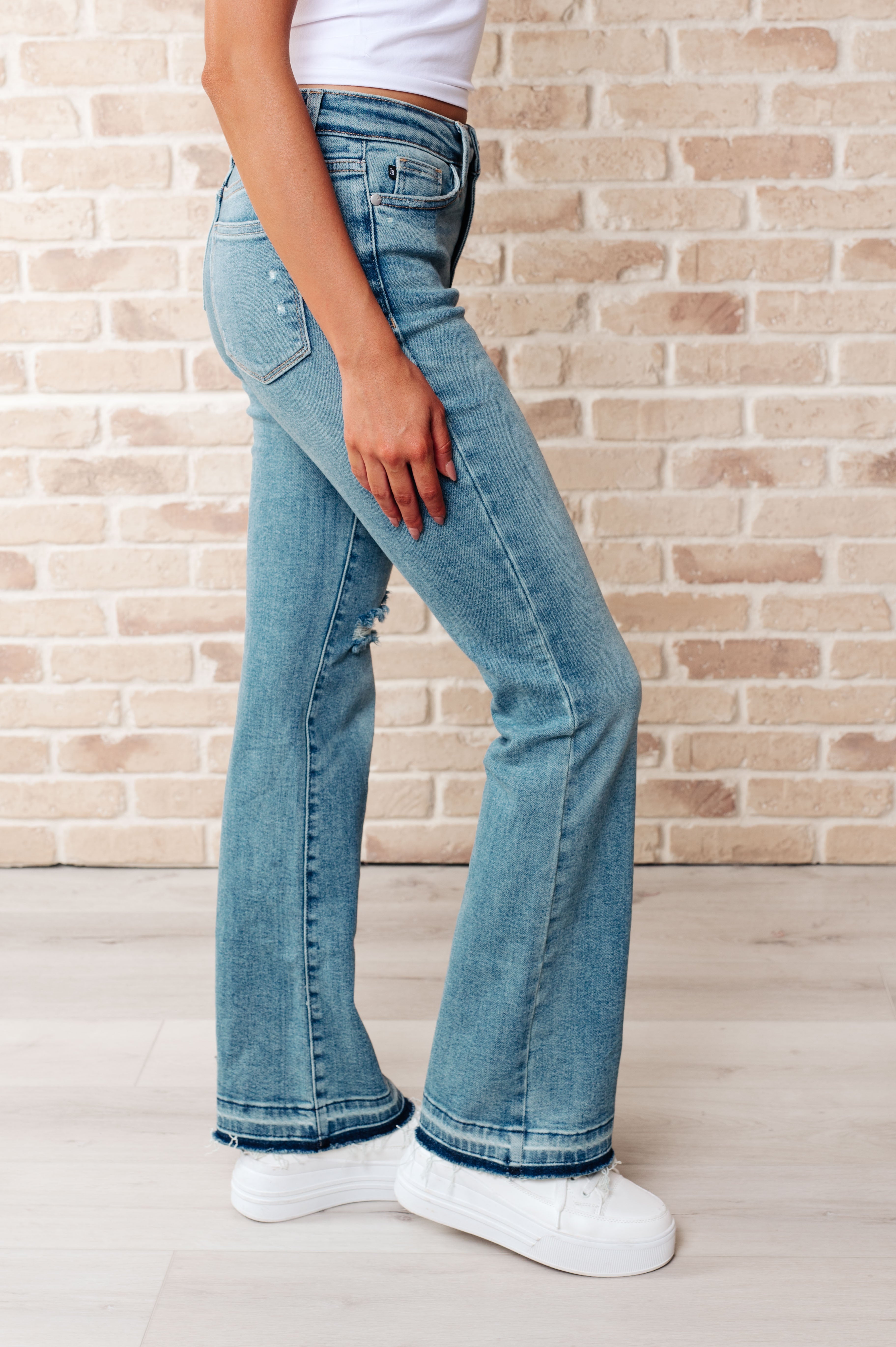 Side profile of person wearing blue Isla mid rise distressed bootcut jeans with released hems and white sneakers indoors