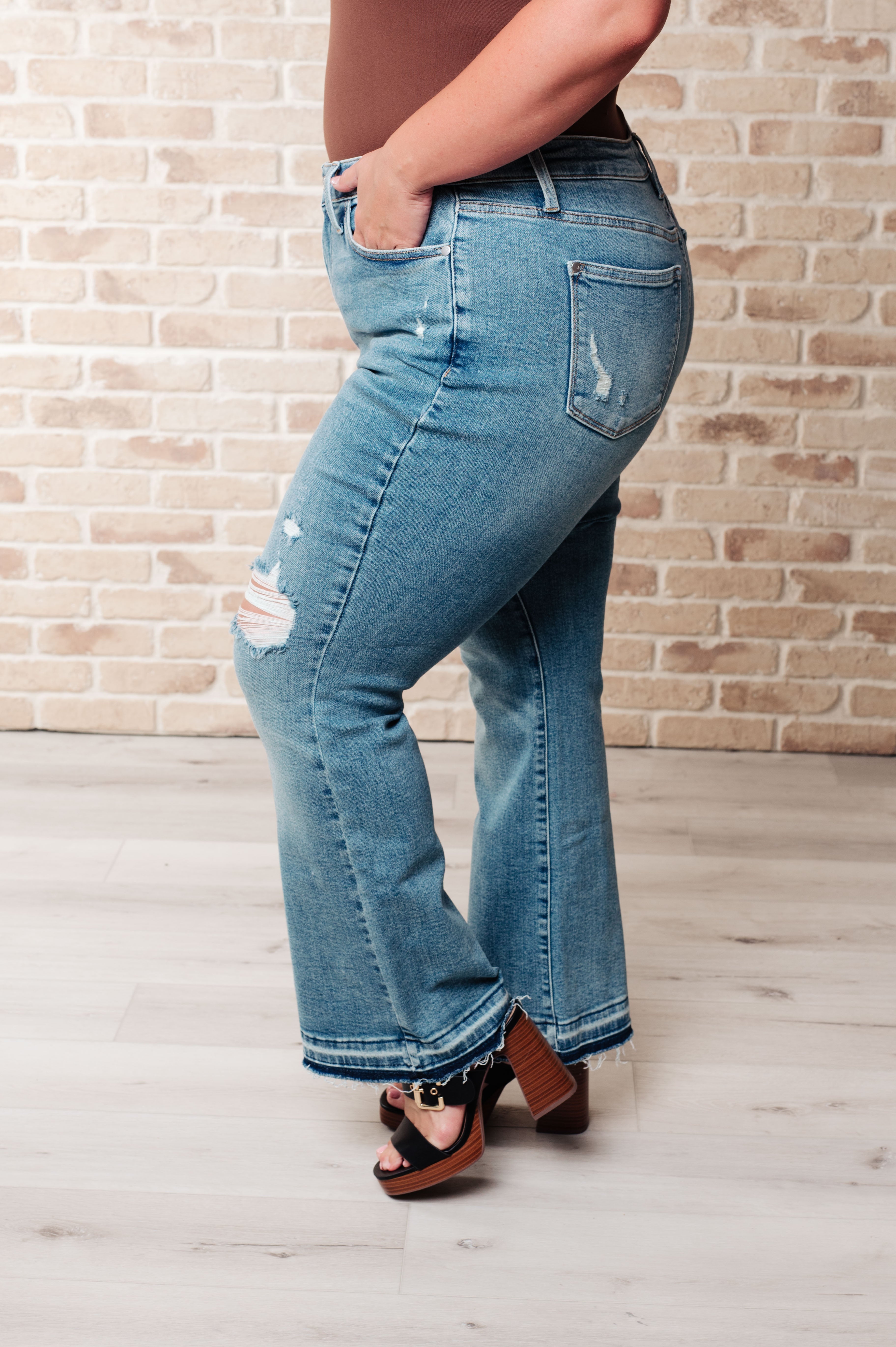 Side view of person wearing blue Isla mid rise bootcut jeans with distressed details and released hems paired with black platform sandals indoors