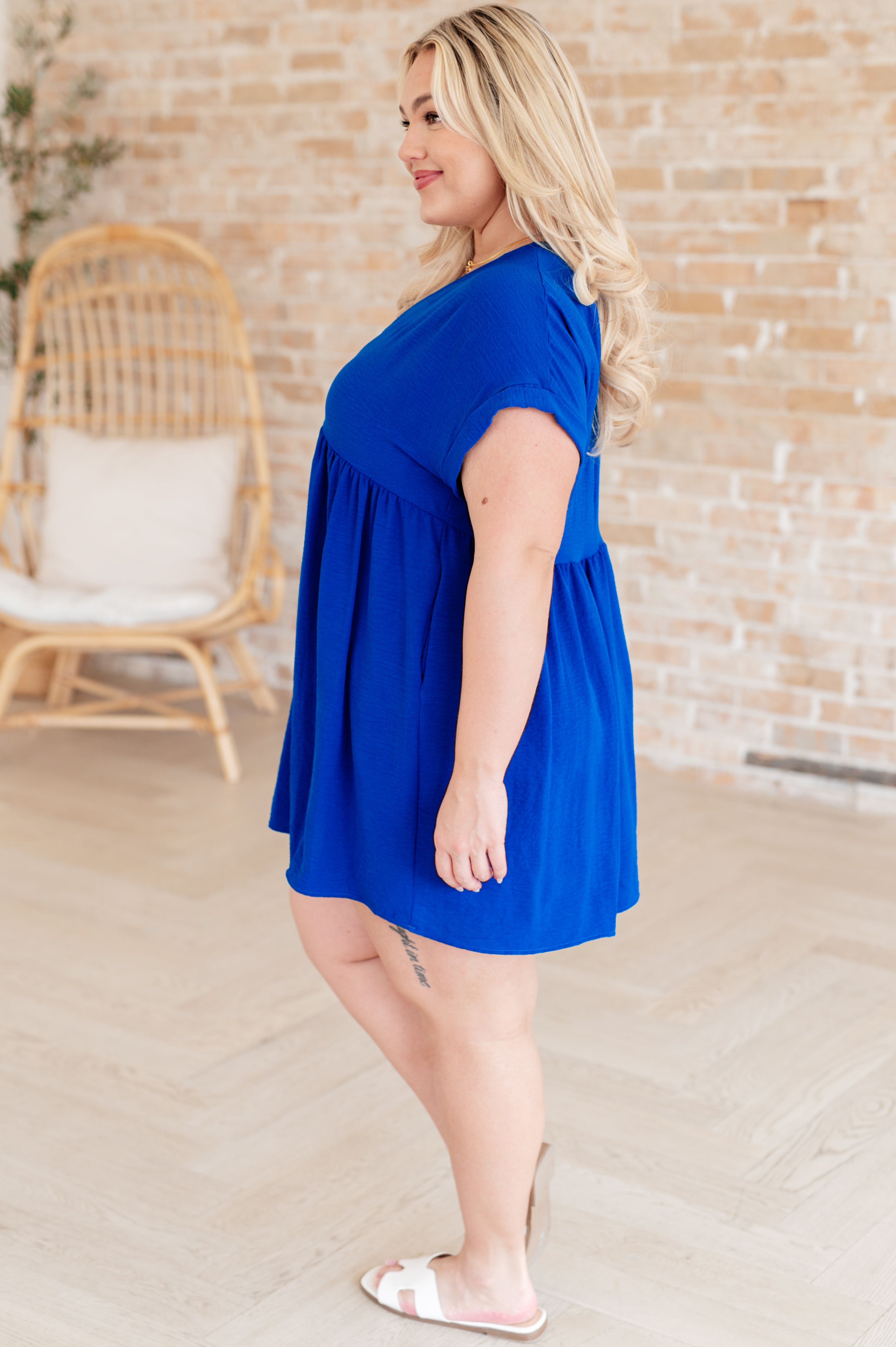 Curvy woman in vibrant blue V-neck dress with short sleeves and relaxed fit paired with white slide sandals indoors