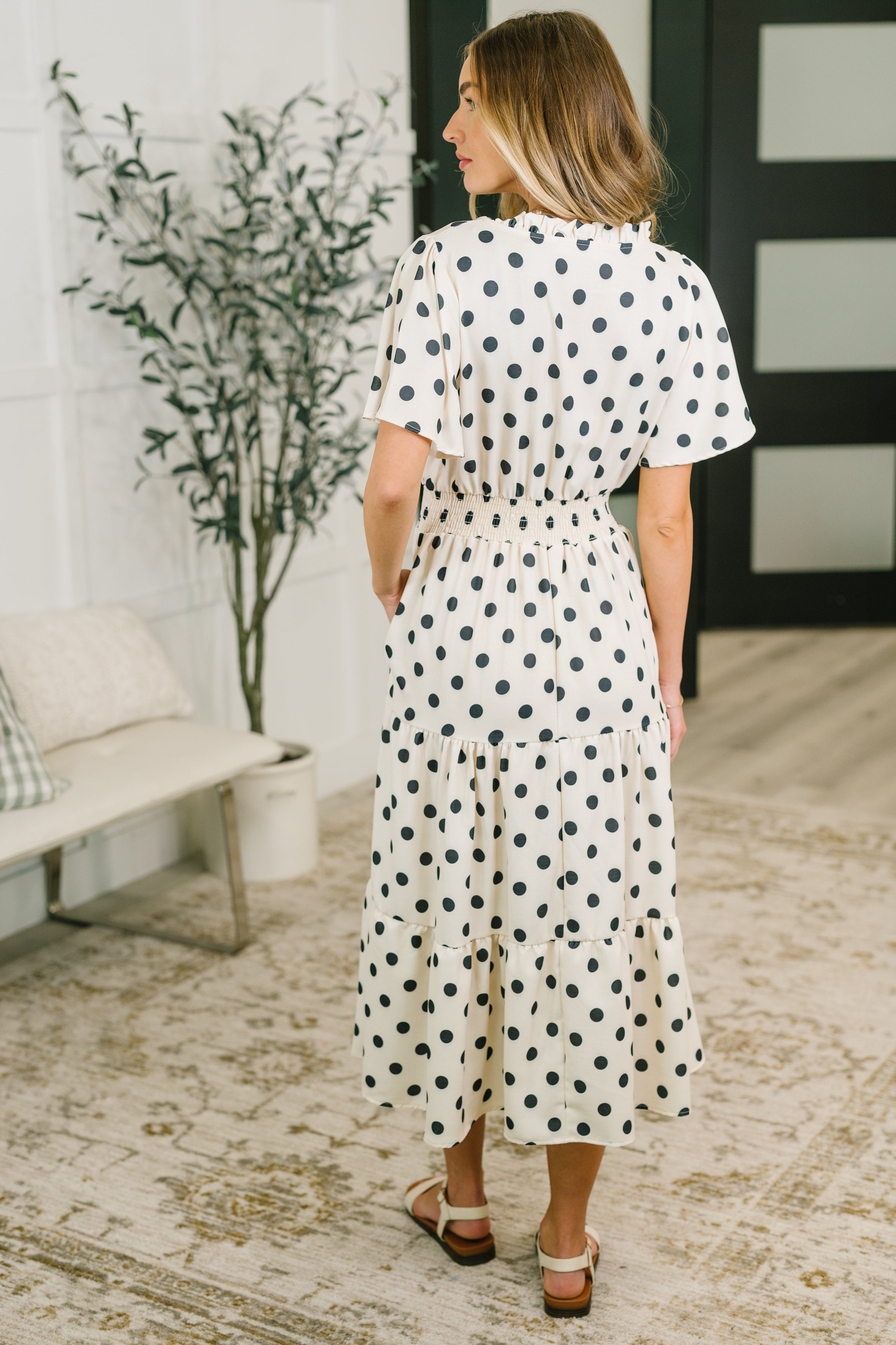 Woman wearing ivory midi dress with black polka dots, gathered waist, and tiered skirt in cozy indoor setting