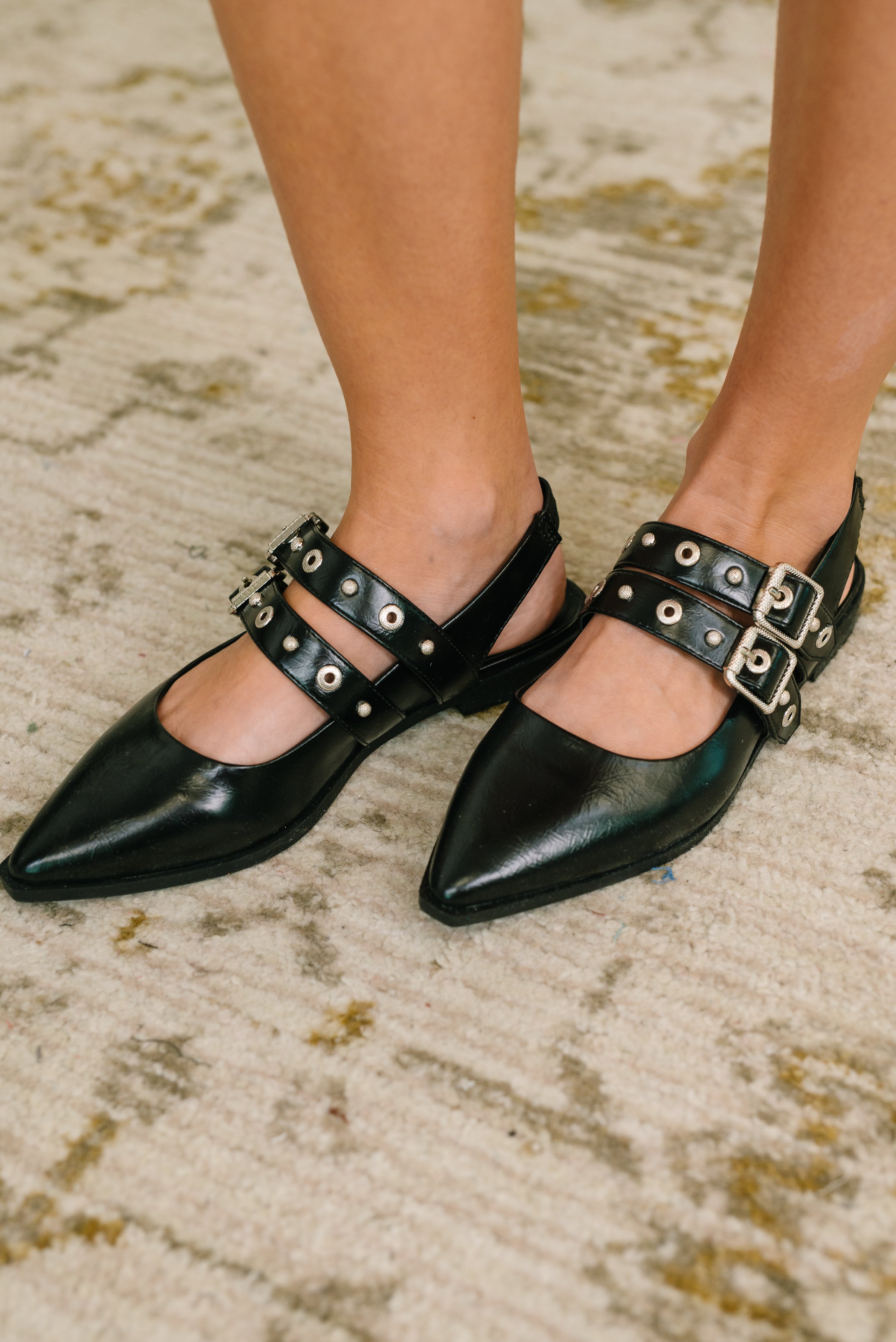 Woman wearing black pointy toe slip-on flats with double buckle straps and silver eyelets on patterned rug