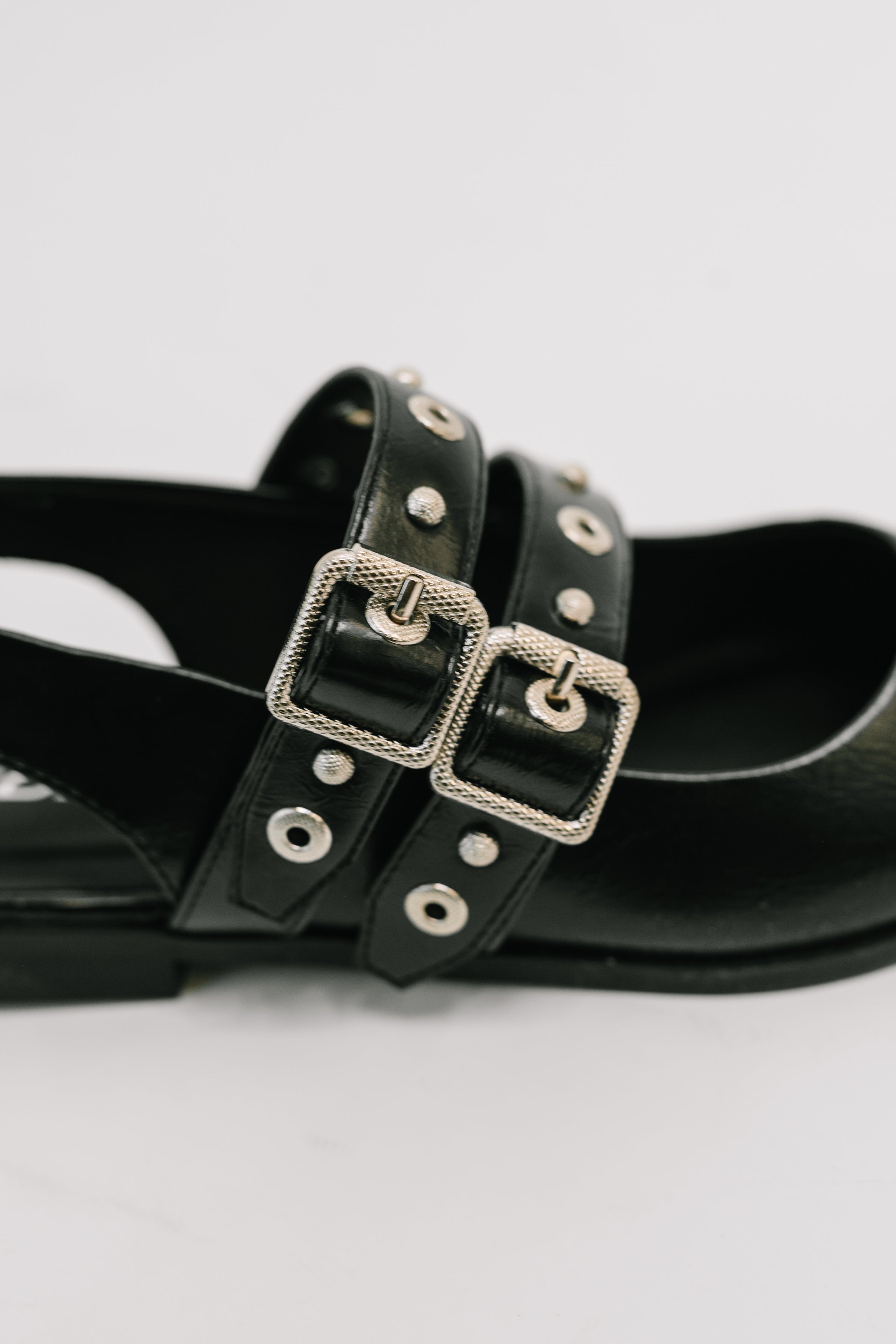 Close-up of black pointed toe slip-on flat featuring two silver buckled straps with stud accents on white background