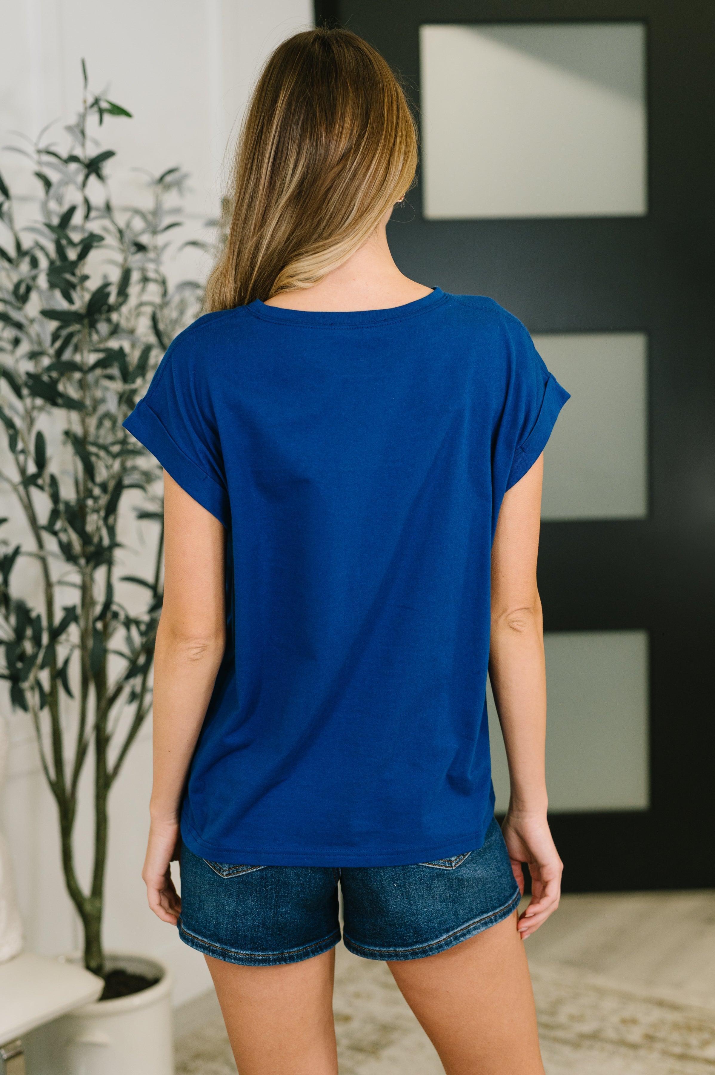 Woman wearing blue basic rolled sleeve tee paired with denim shorts standing indoors with back facing camera