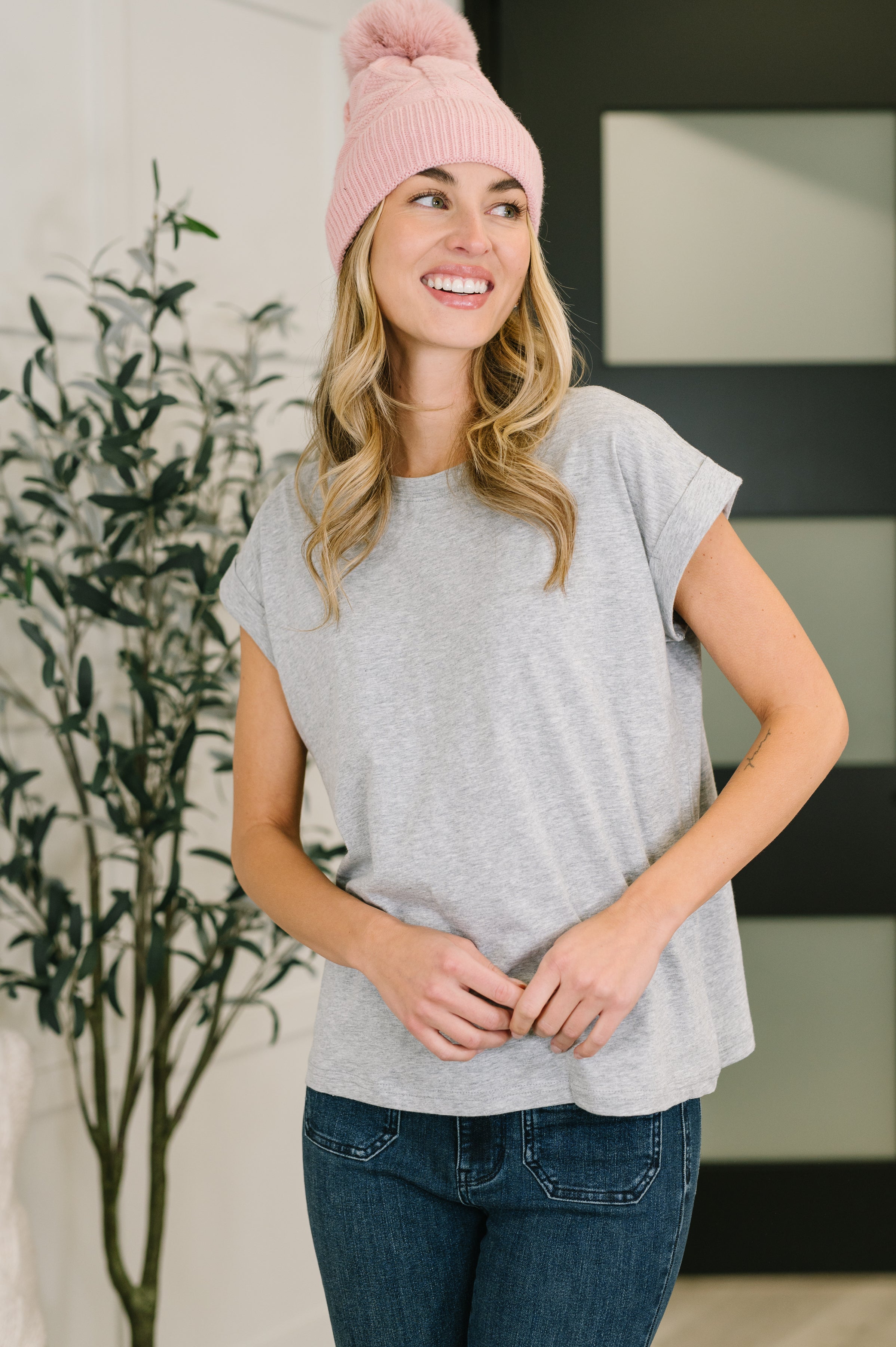 Smiling woman wearing grey rolled sleeve tee and pink knit beanie standing indoors with hands clasped in front