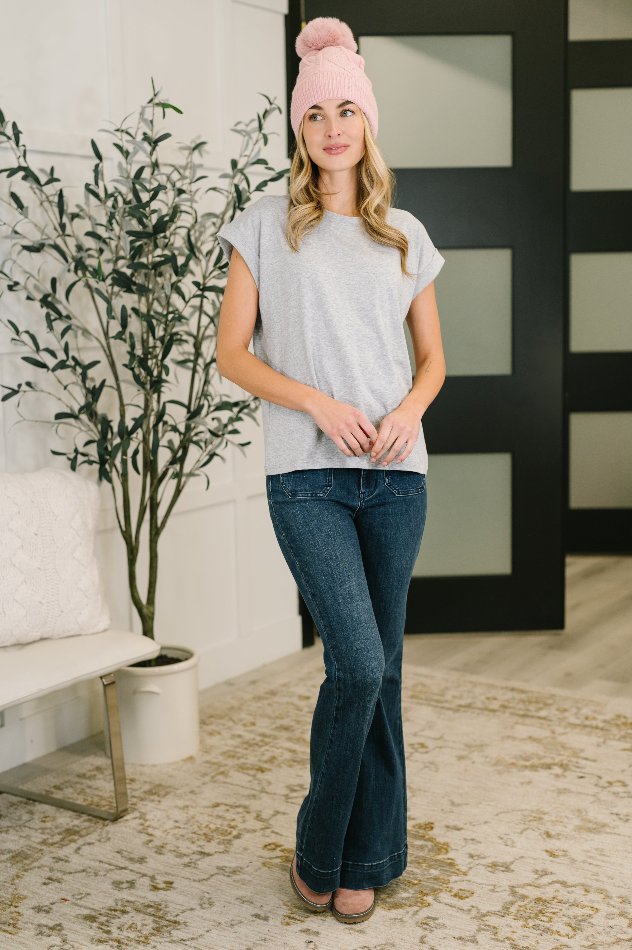 Woman wearing grey rolled sleeve tee with dark bootcut jeans and pink pom-pom beanie standing indoors