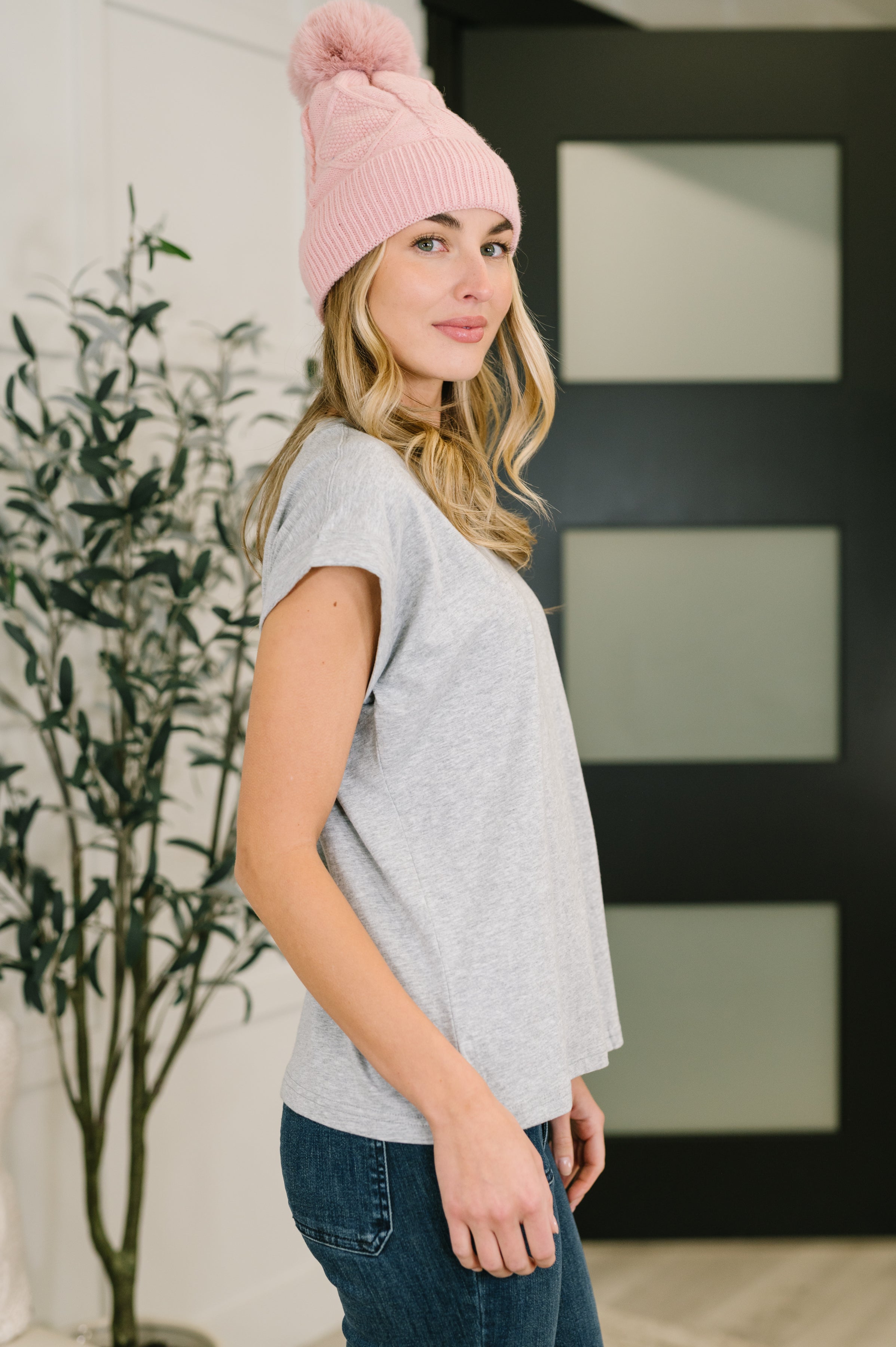 Woman wearing a grey basic rolled sleeve tee with pink knit pom-pom beanie and blue jeans standing indoors in side profile