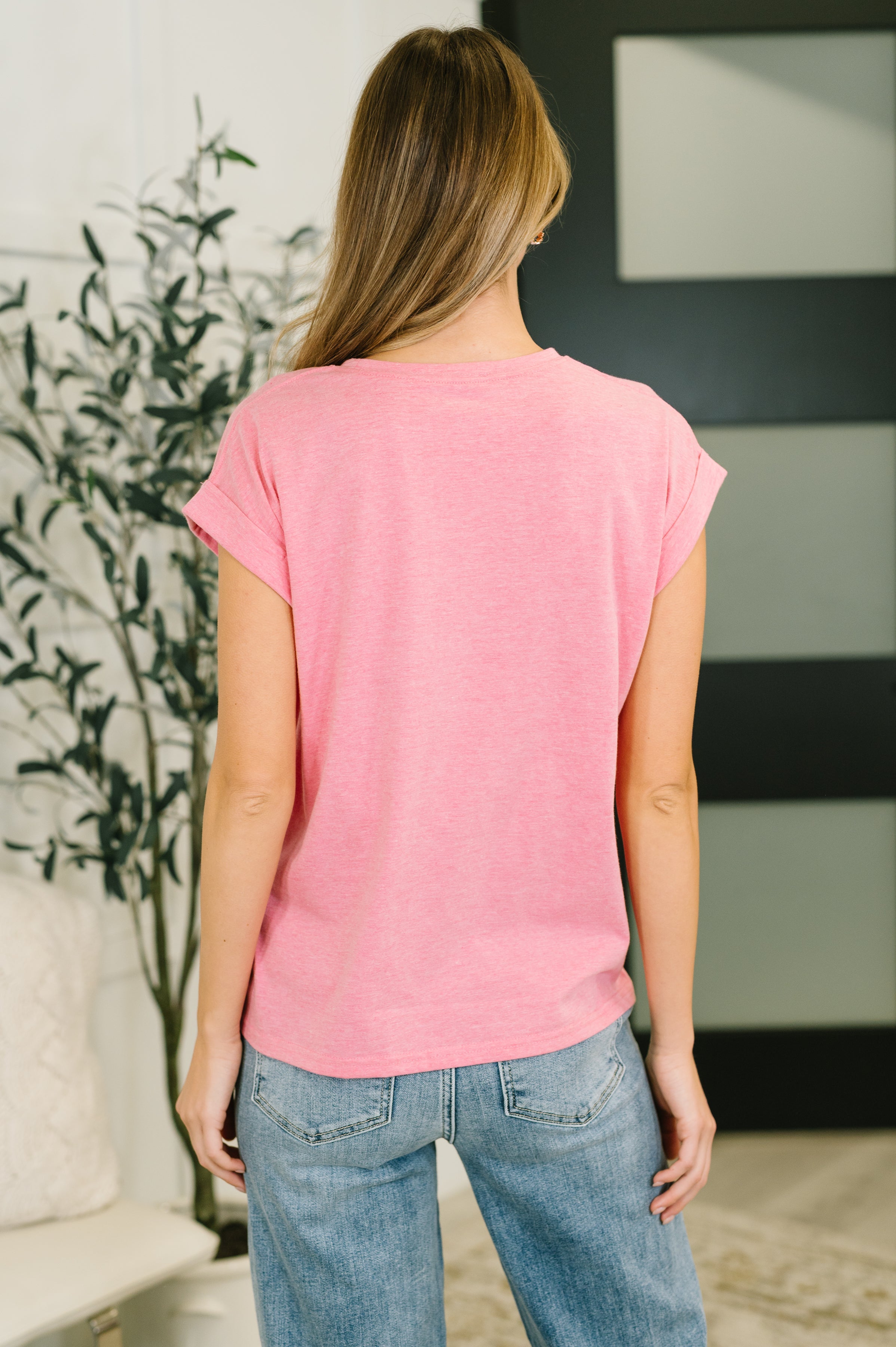 Woman viewed from back wearing pink rolled sleeve tee and light blue jeans standing indoors near plant and door
