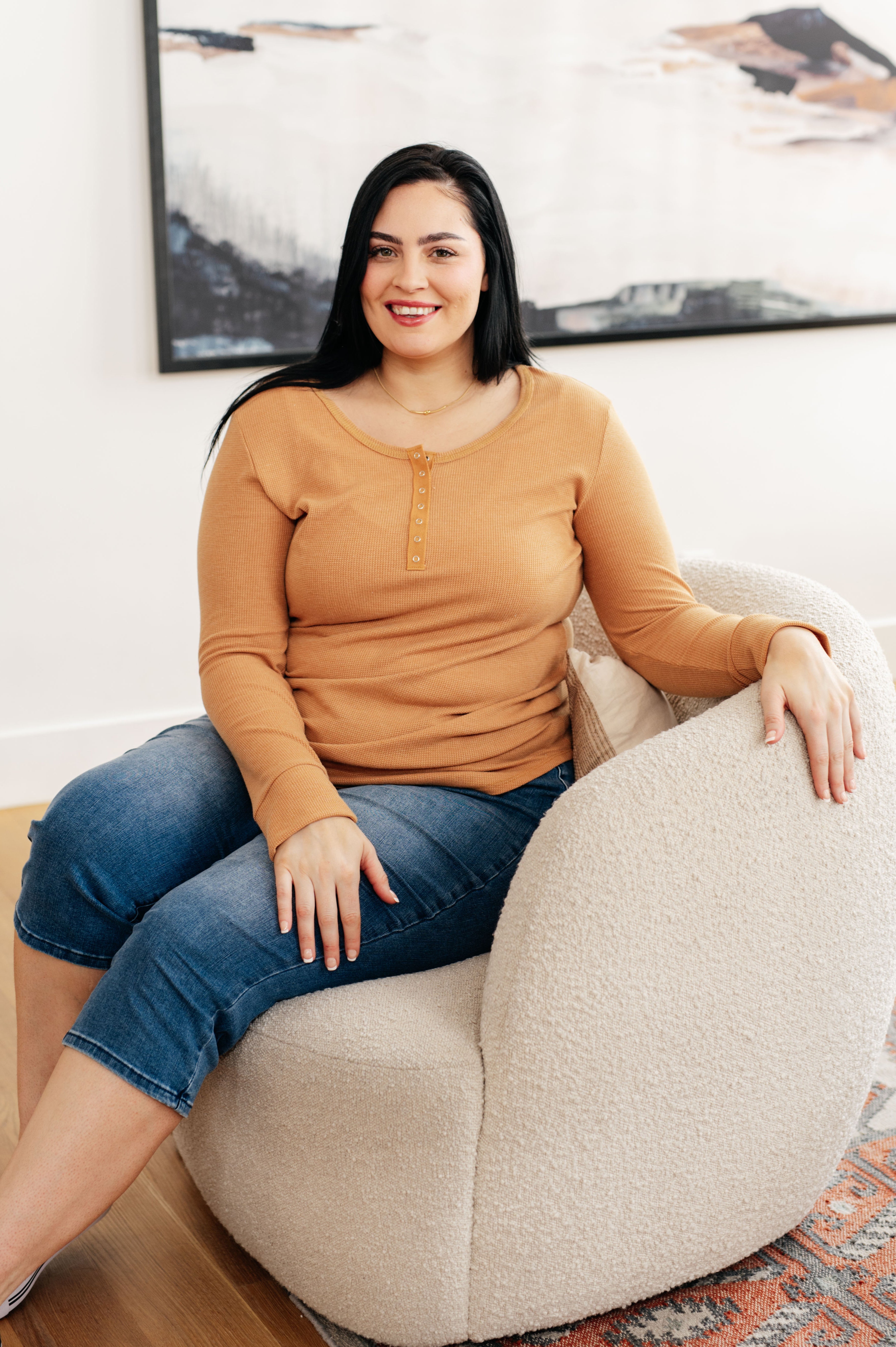 Person sitting on cream textured chair wearing golden sand long sleeve Henley top with snap button placket and cropped blue jeans indoors