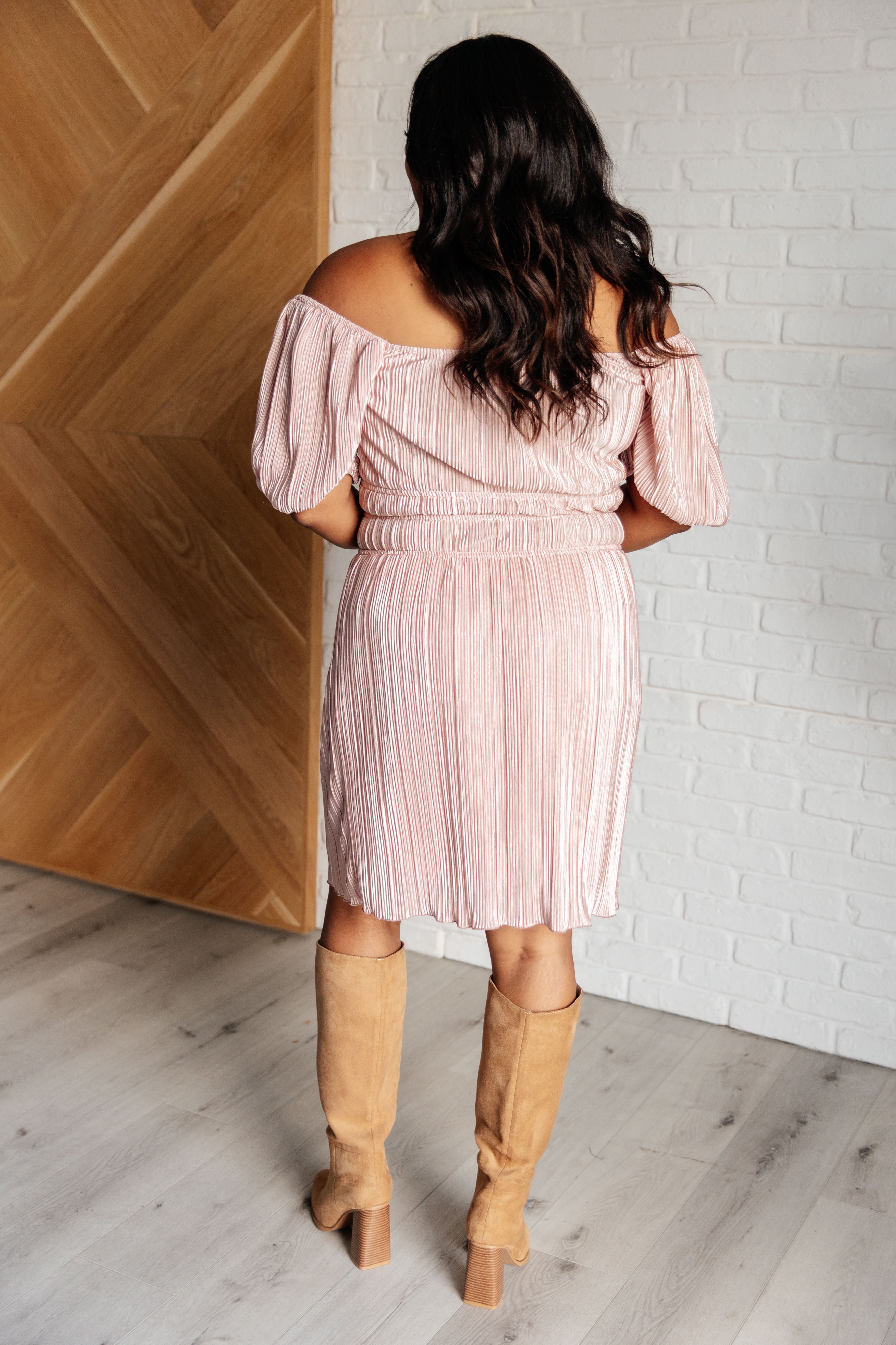 Woman wearing a pink off-shoulder pleated dress with puffed sleeves paired with tan suede knee-high boots indoors