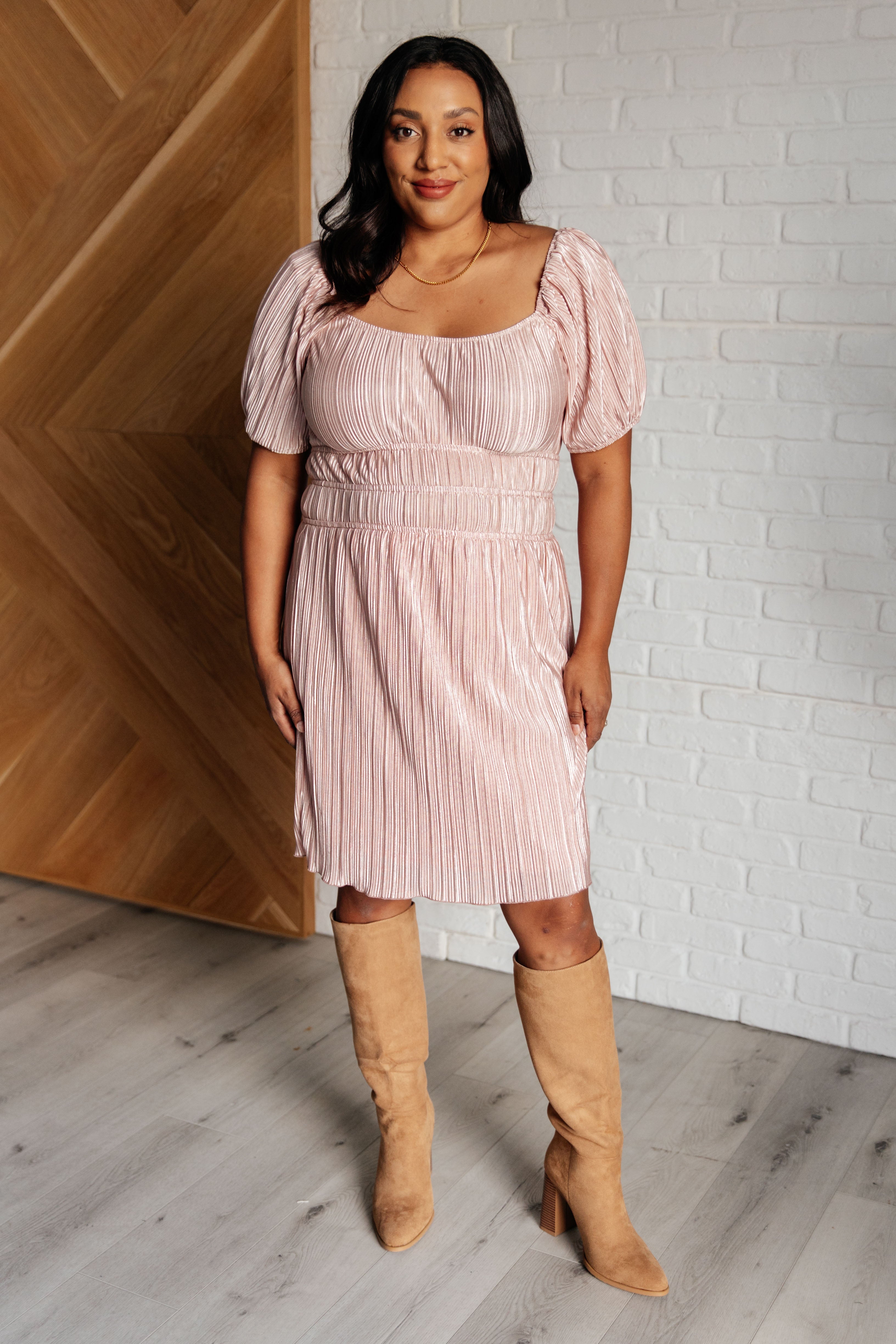 Woman in a pink pleated dress with puffed short sleeves and square neckline wearing tan suede knee-high boots indoors