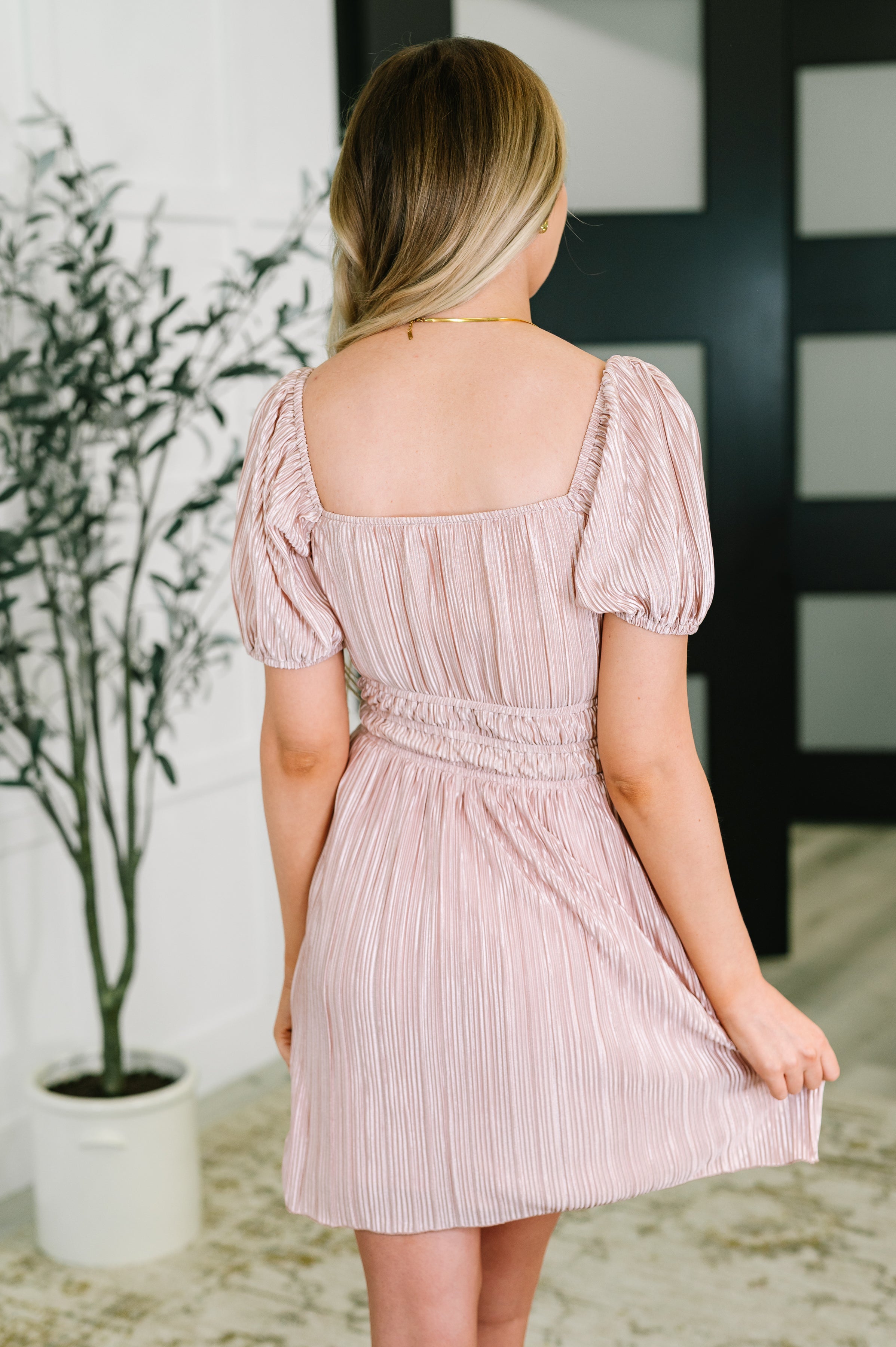 Woman wearing a soft pink dress with gathered waist and puffed short sleeves shown from back indoors