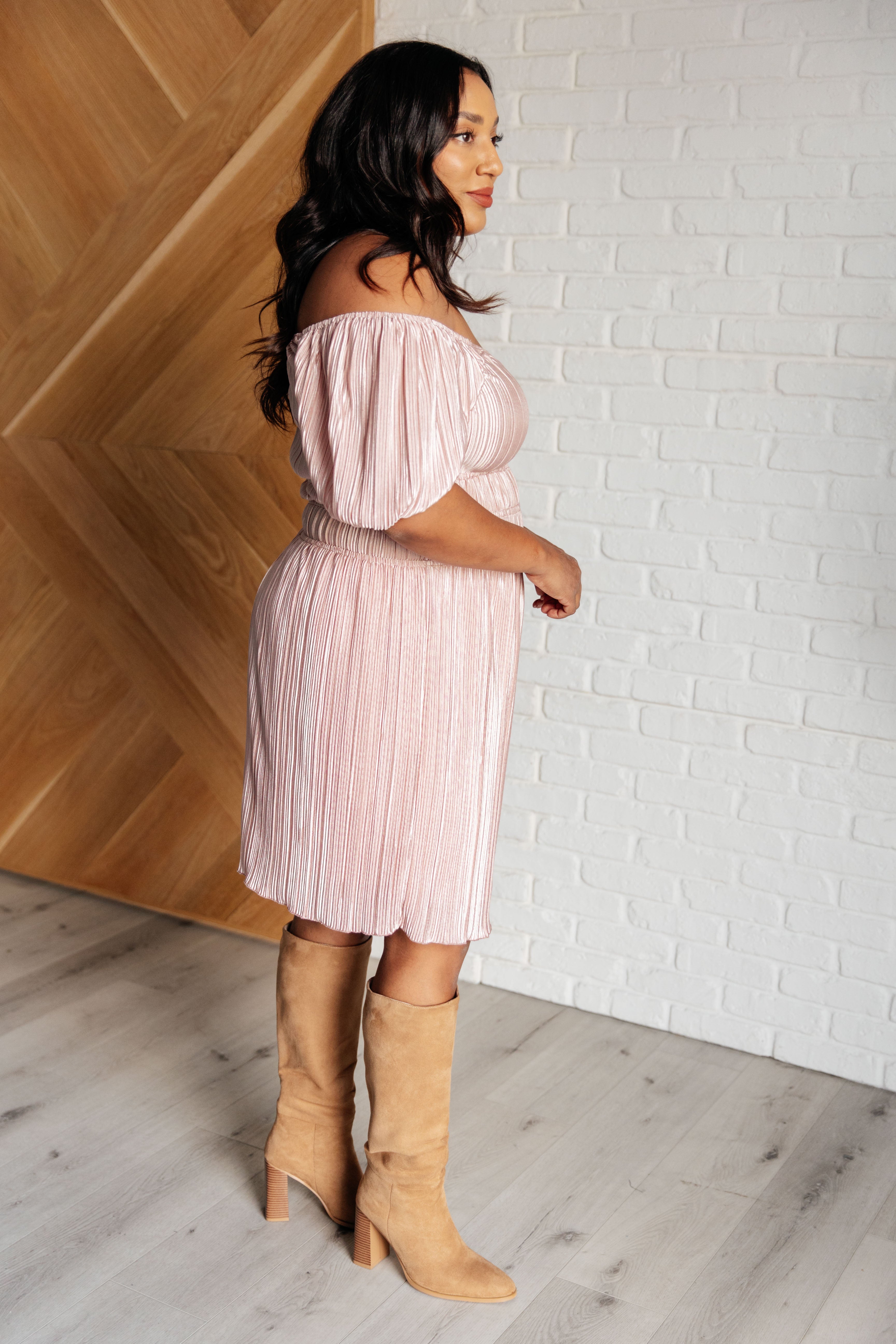 Woman wearing a light pink off-shoulder dress with balloon sleeves and pleated skirt paired with tan suede knee-high boots indoors