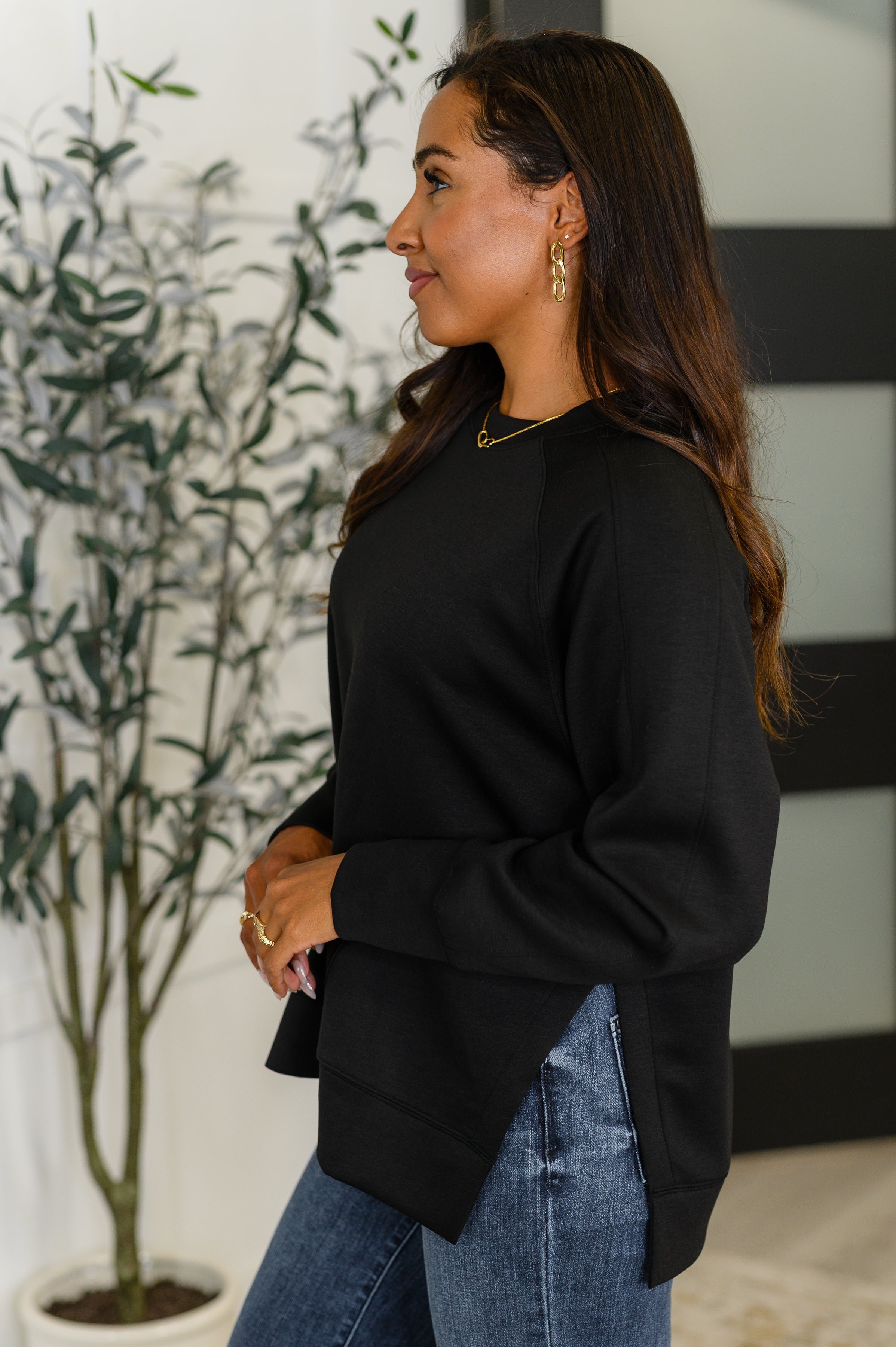 Profile of woman wearing black relaxed scuba pullover with side slits paired with blue jeans indoors next to plant